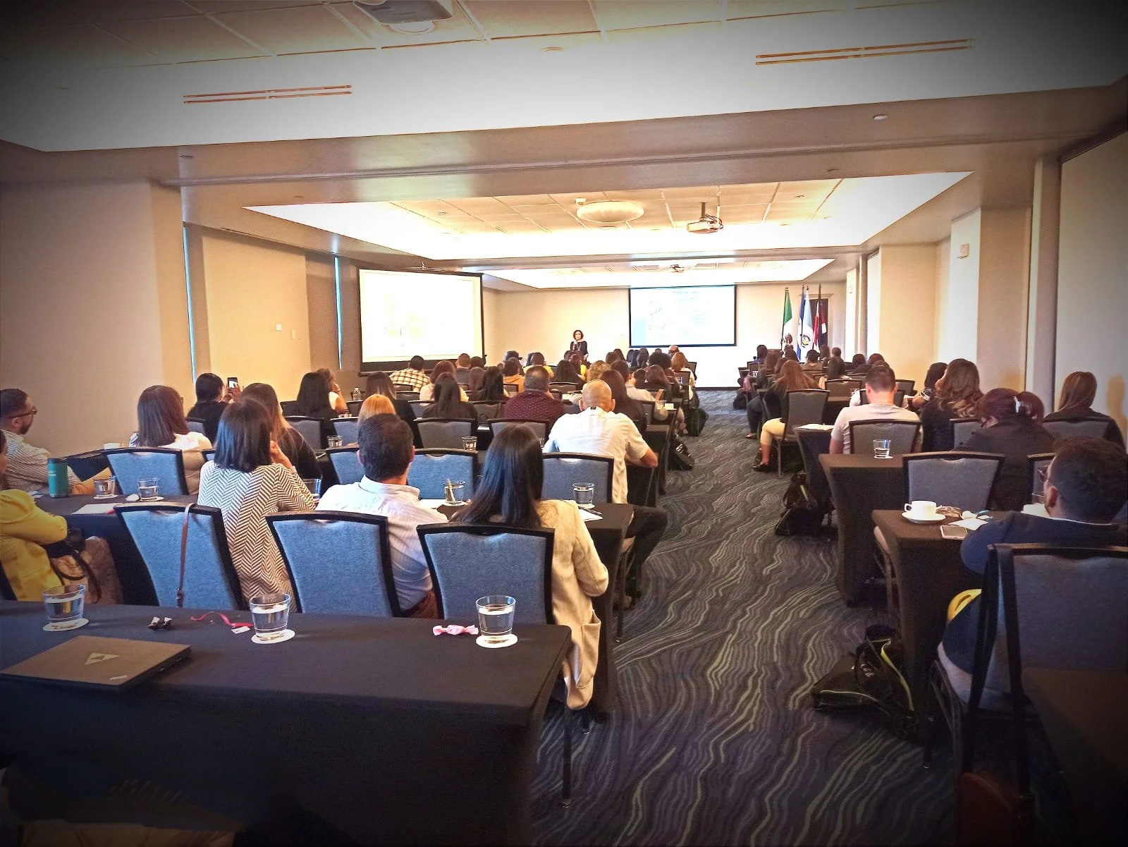 Auditorio lleno de personas en una conferencia, con una presentadora en el escenario y dos pantallas grandes detrás, en un ambiente formal con banderas en el fondo.