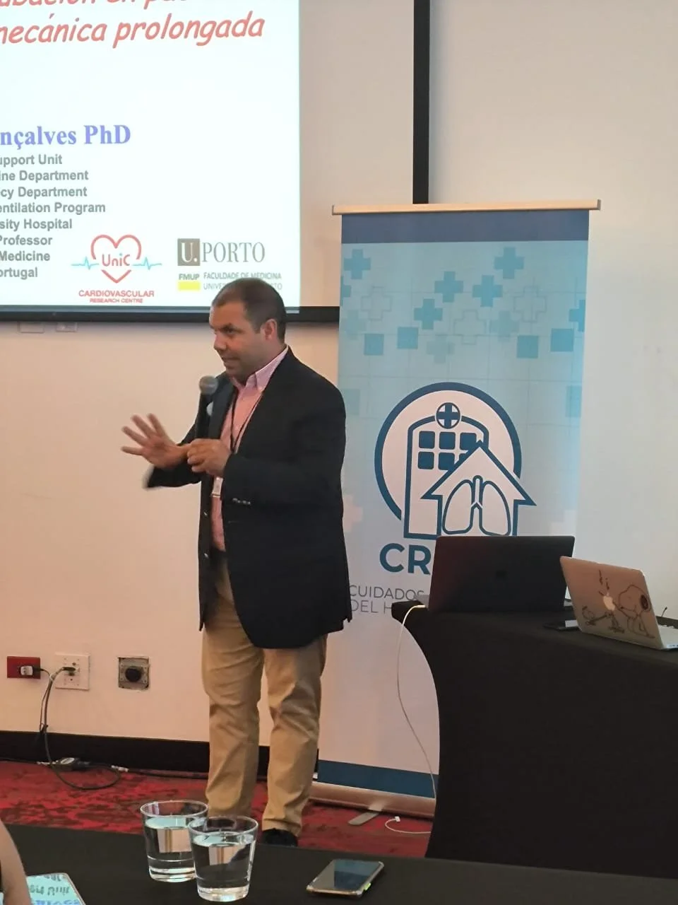Hombre dando una conferencia en un evento de medicina, con pantalla y banner relacionados con salud y cuidados del pulmón.