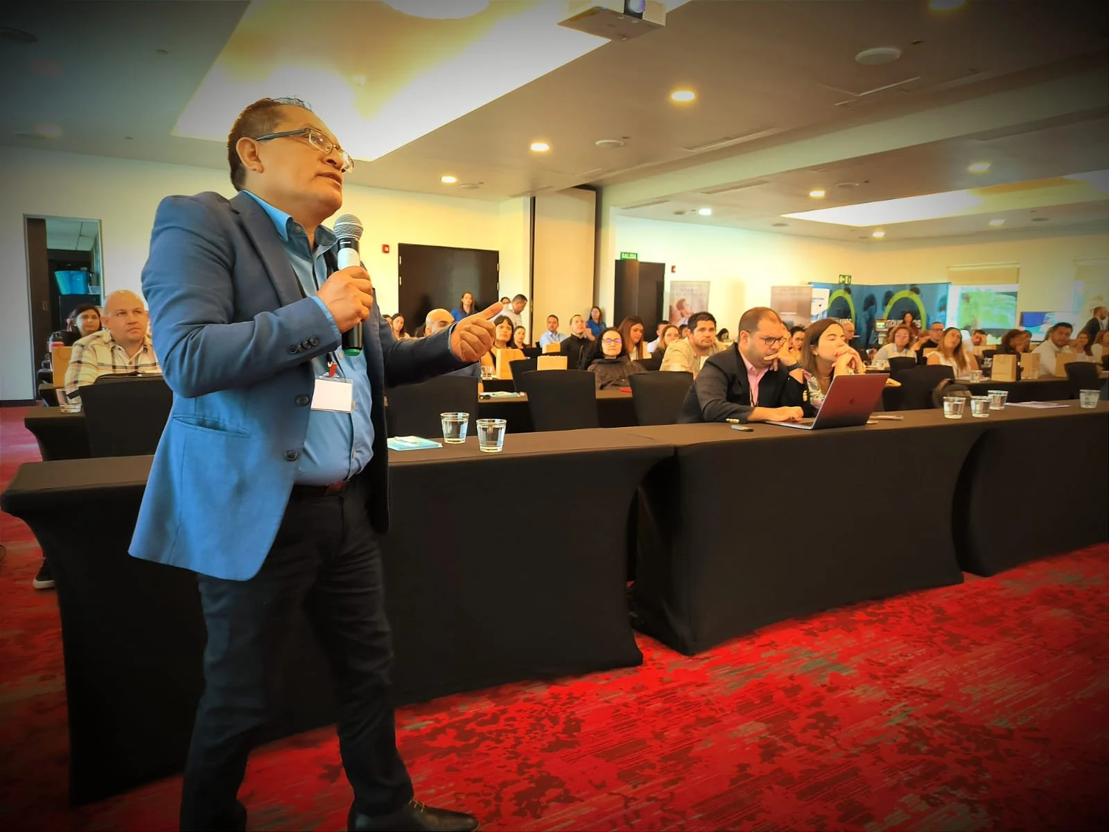 Hombre de pie con un micrófono en un evento o conferencia, dando una presentación frente a una audiencia en una sala de conferencias.