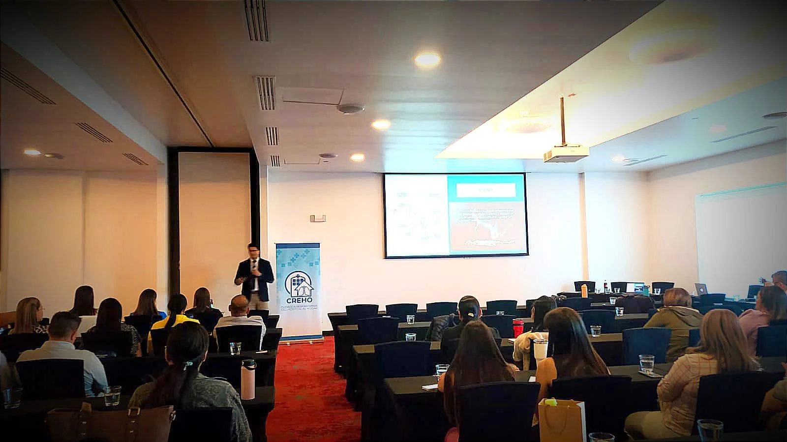 Conferencia en un salón con muchas personas, un presentador en el escenario y una pantalla grande mostrando una diapositiva.