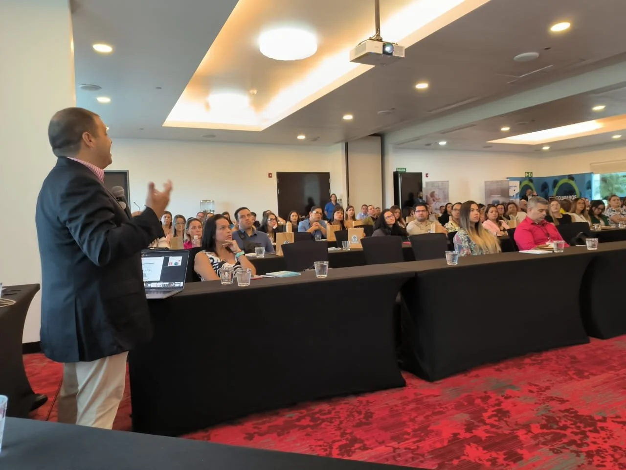 Un orador vestido con saco negro y camisa de rayas, presentando frente a una audiencia en una conferencia en un salón con cielo raso iluminado.