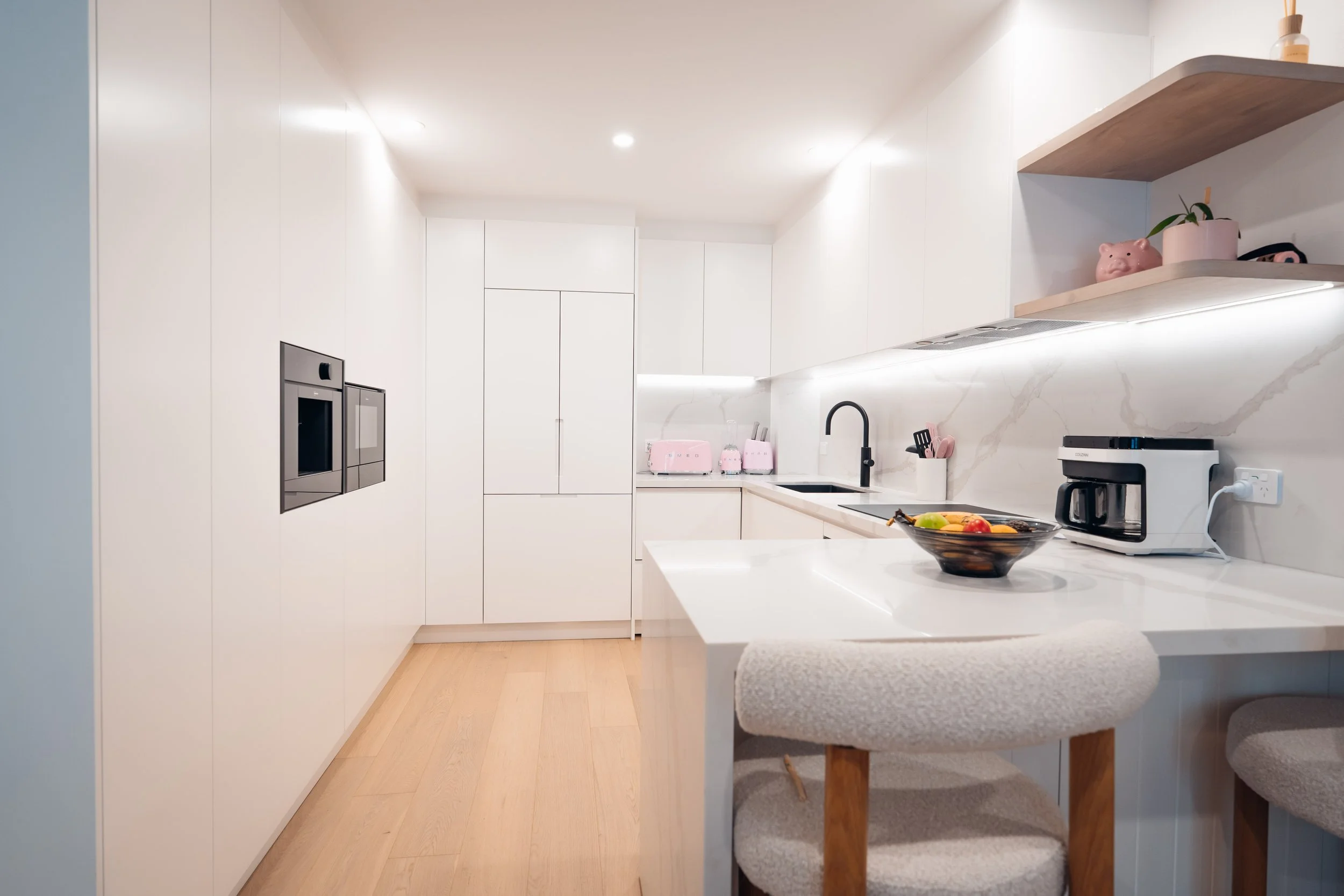 Modern white kitchen with built-in appliances, wooden shelves, and light wood flooring.