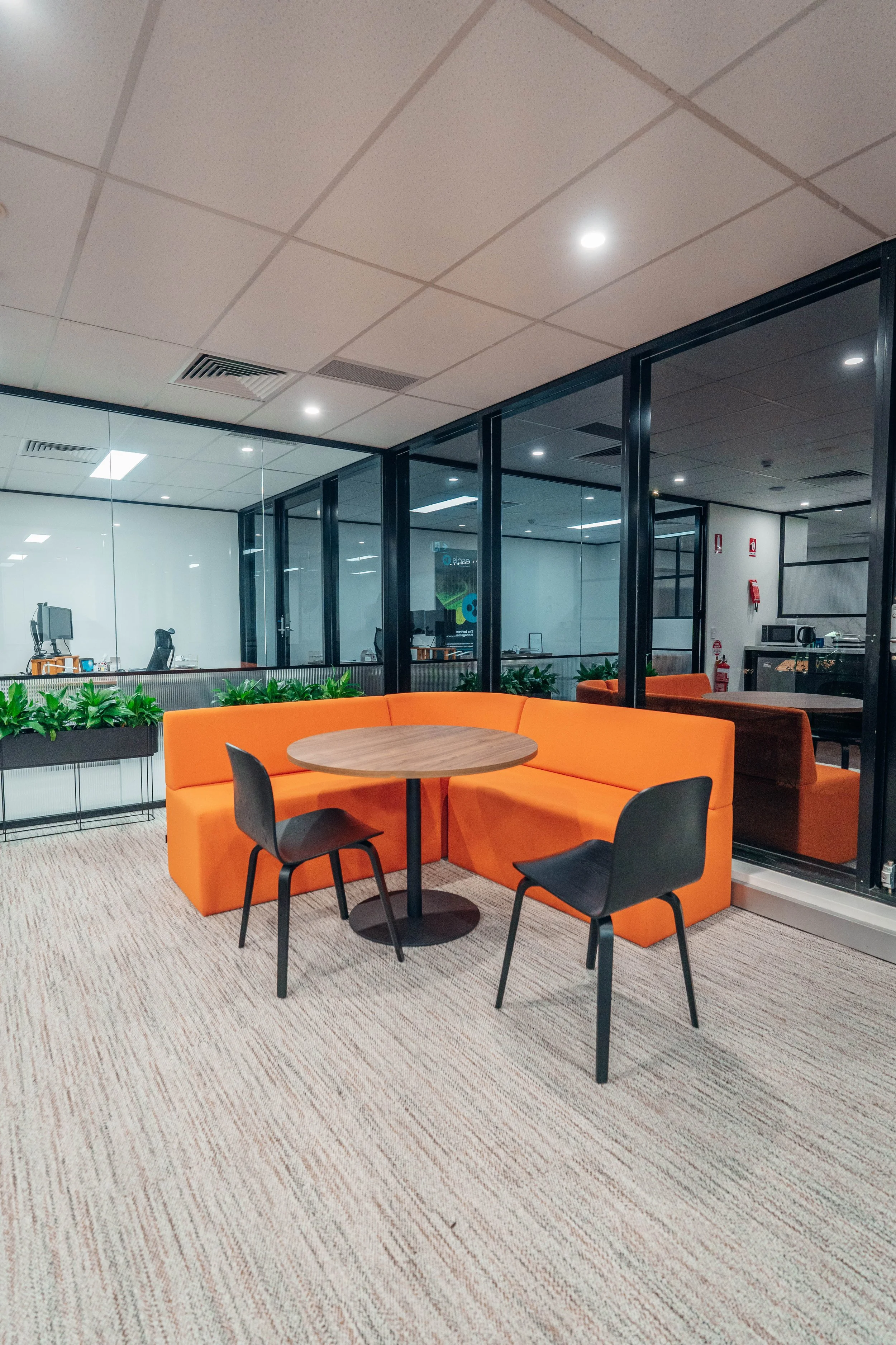An office lounge area with an orange cushioned sectional sofa, a round wooden table, and two black chairs on a beige carpet. Behind the seating, there is a glass wall with an office space visible through it, with desks, computers, and office chairs. The ceiling has white tiles with recessed lighting.