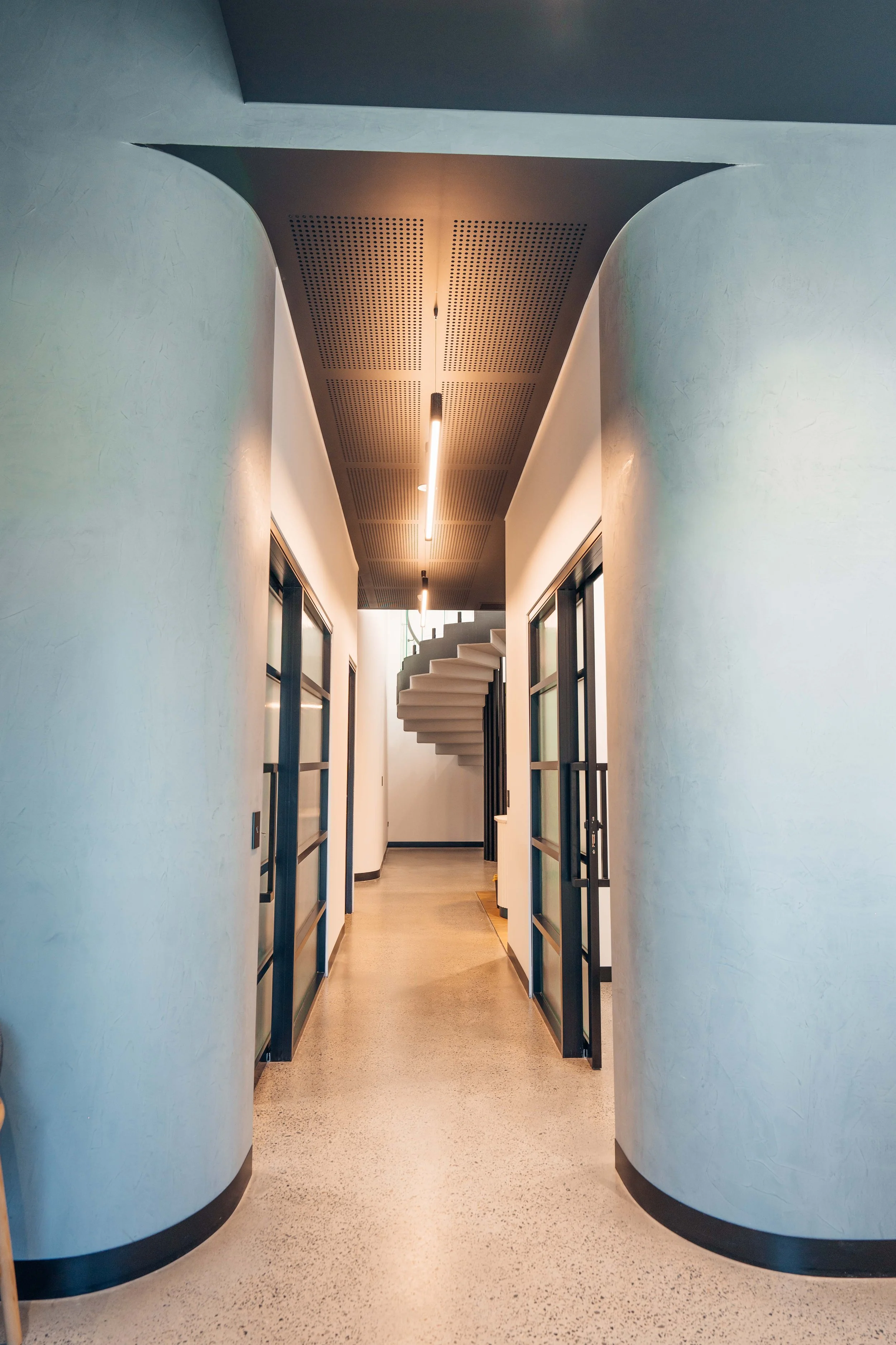 Modern hallway with curved walls and glass doors, leading to a spiral staircase at the end, illuminated by ceiling lights.
