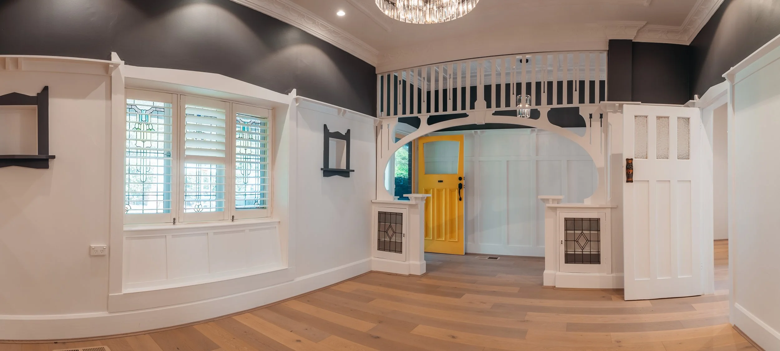 Interior view of a room with white walls, a yellow front door, large windows with white shutters, a dark ceiling with crown molding, a chandelier, and a loft area with a white railing.