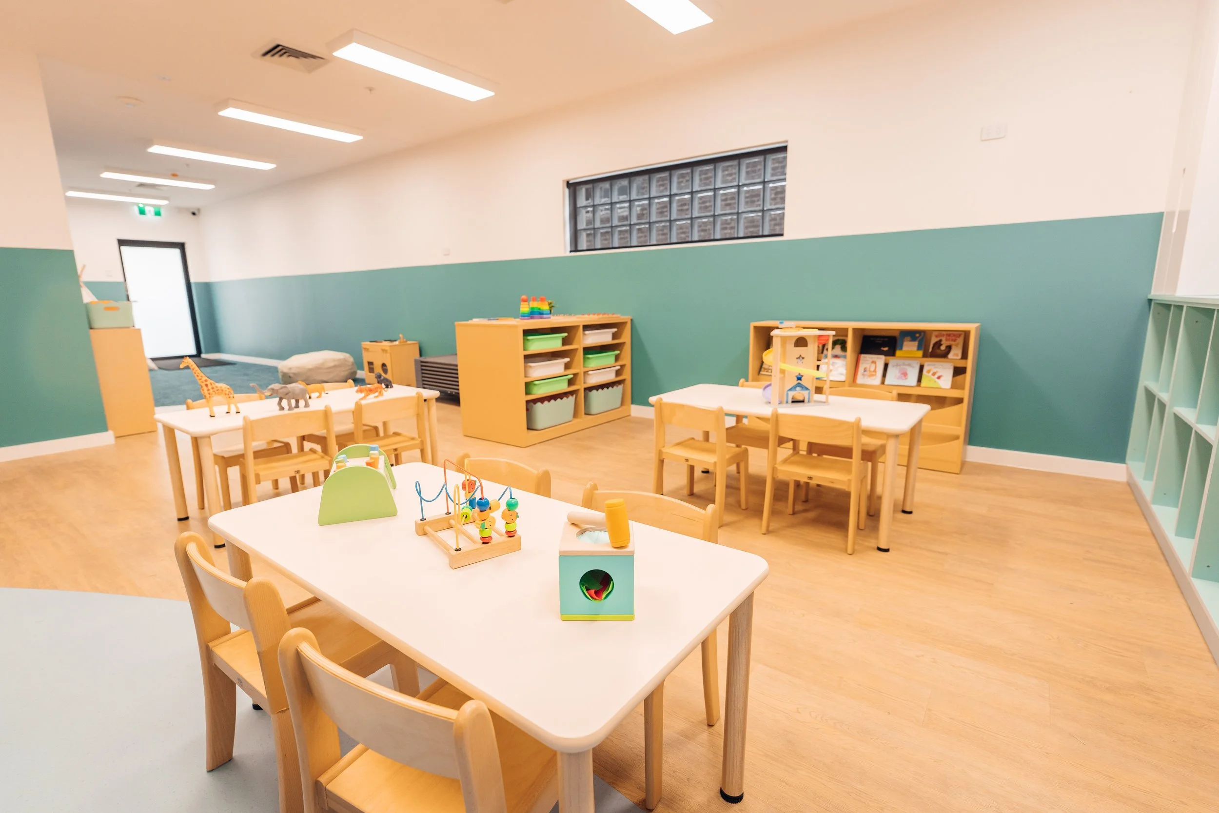 Bright preschool classroom with wooden tables and chairs, stuffed animal toys, art supplies, and books on shelves, with a green and white color scheme and natural light.