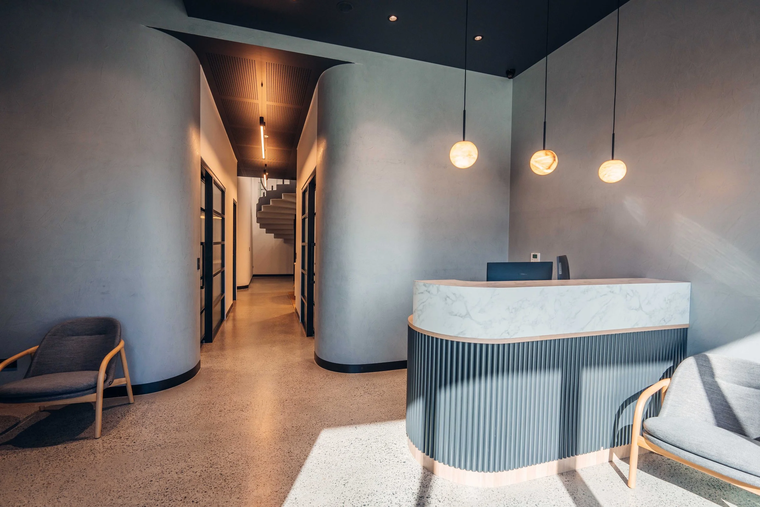 Modern reception area with a curved marble desk, pendant lights, and armchairs near the wall, leading to a hallway with a spiral staircase.