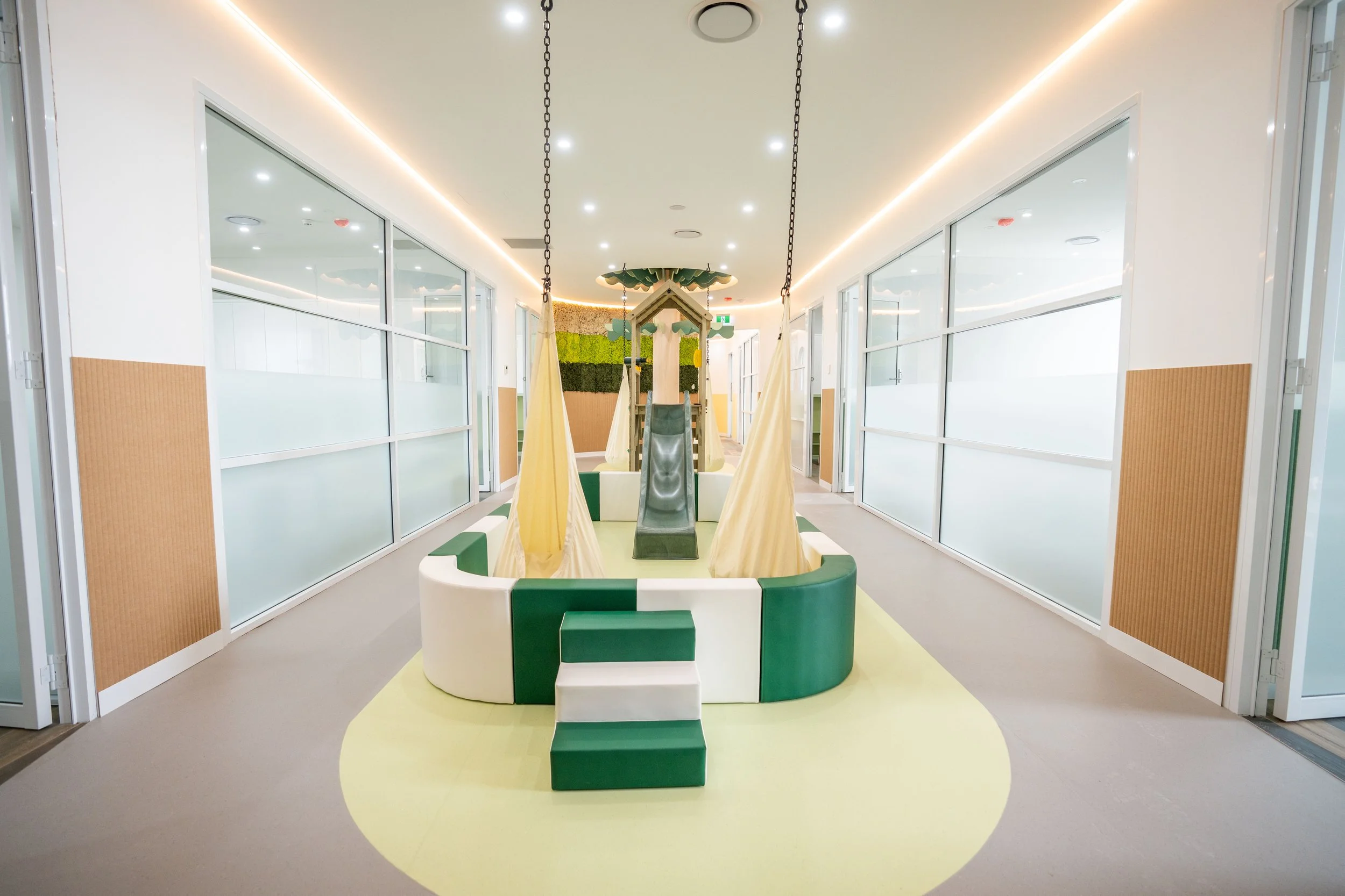 Indoor children's play area with a small slide, hanging fabric swings, and padded seating, surrounded by glass walls with frosted sections.