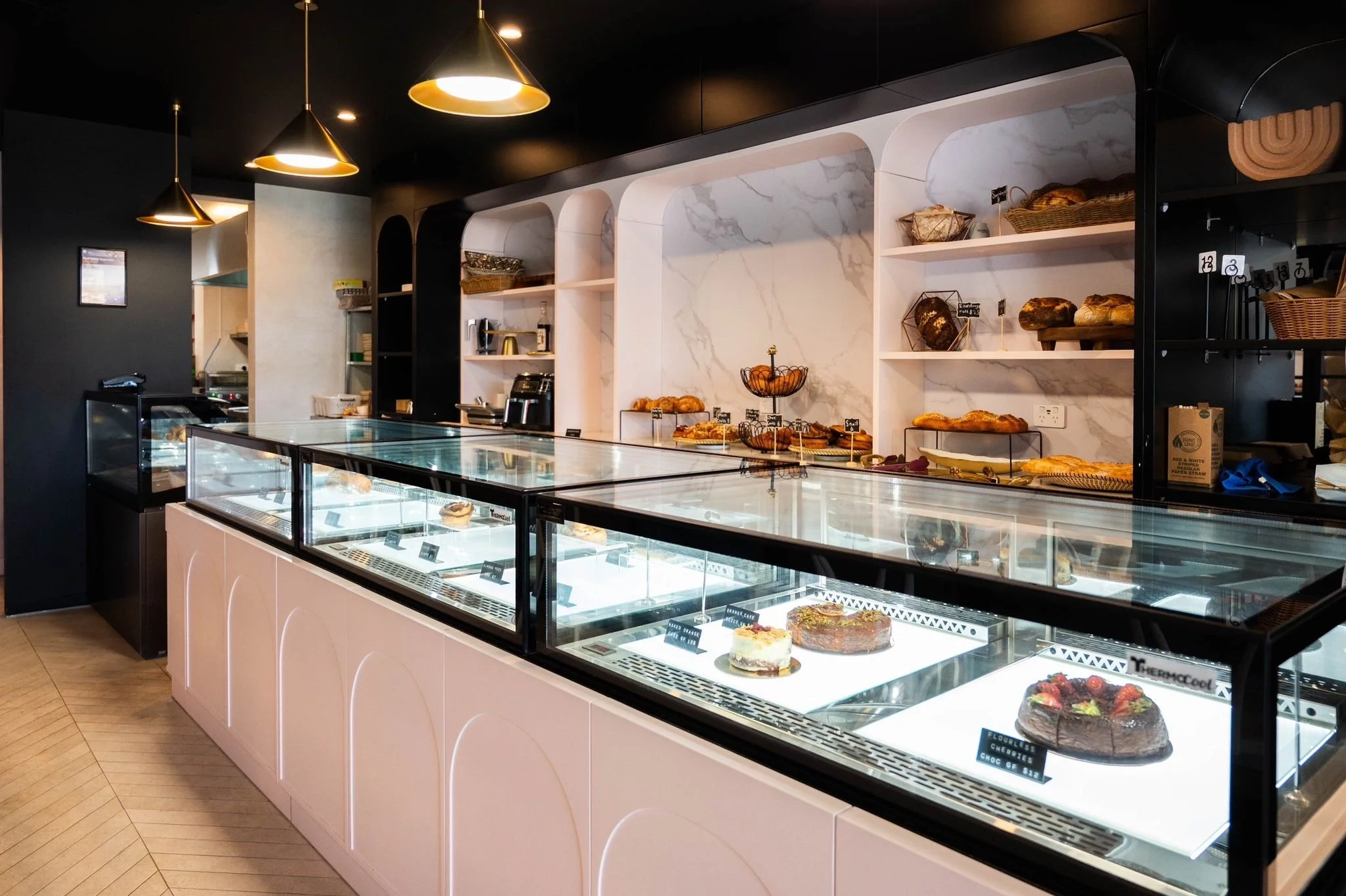 A bakery display case with glass fronts showcasing various cakes and pastries. Shelves behind the case hold bread and baked goods, with black, white, and marble decor and warm lighting.