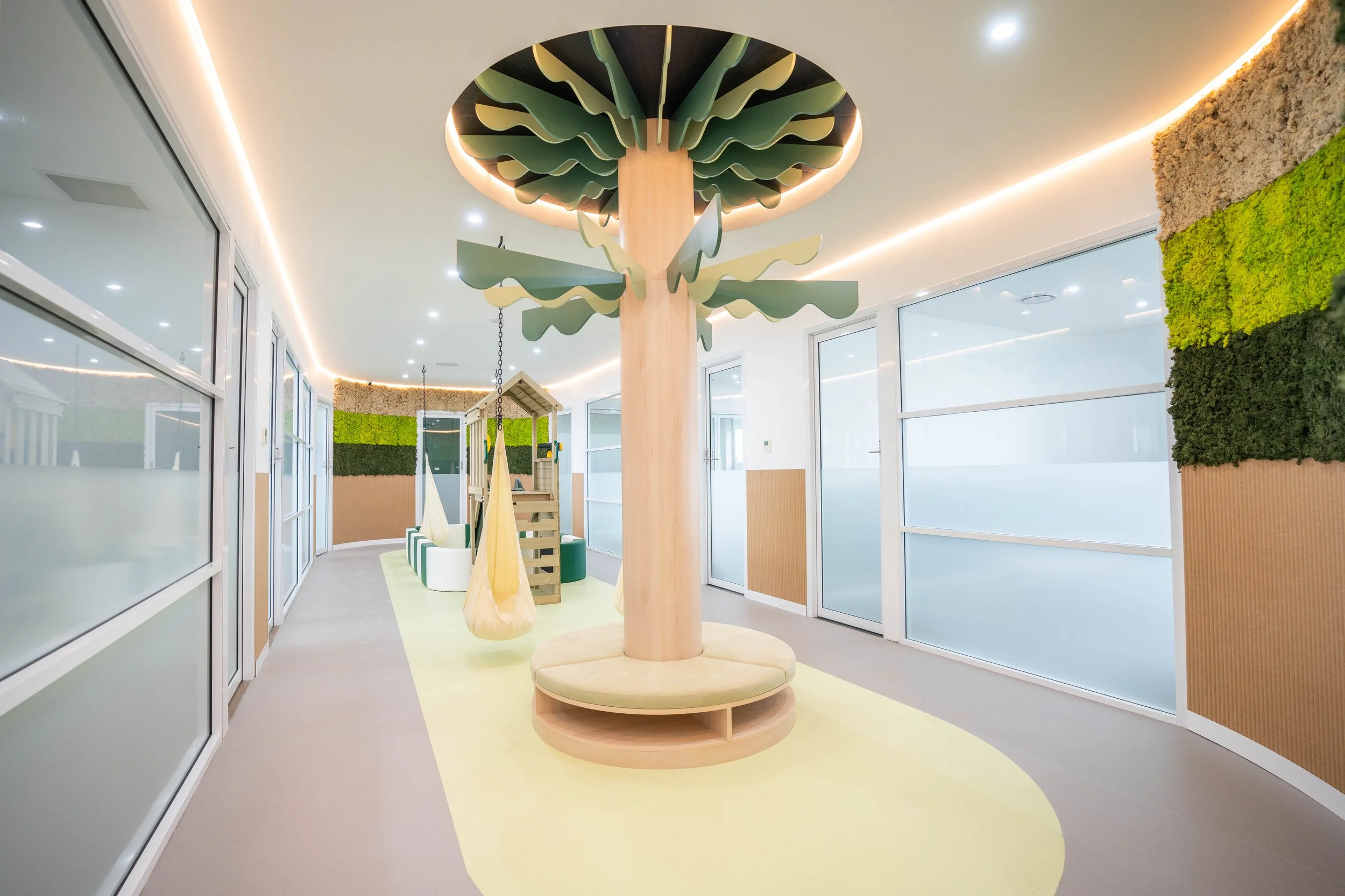 Interior of a child-friendly play area with a large wooden tree structure, hanging fabric swings, and a small playhouse, with decorated walls and large windows.