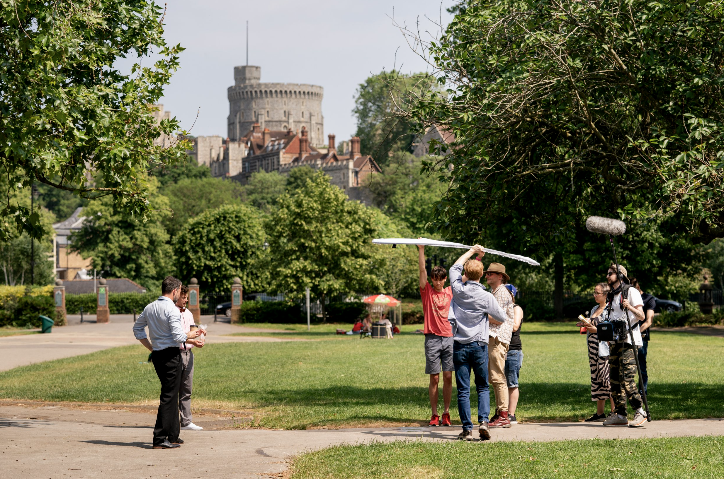 Filming crew and actors on a park, with a historic castle in the background, preparing for a scene.