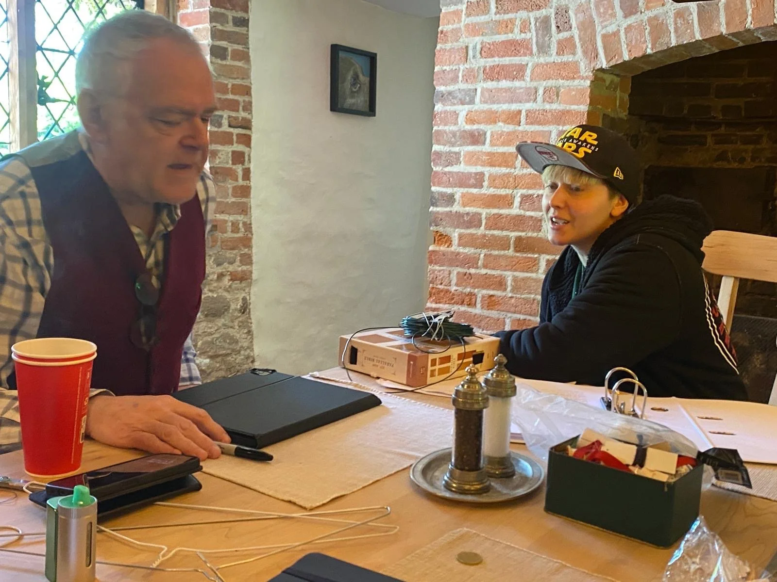 Two men sitting at a wooden table inside a room with brick and white walls, having a conversation. The man on the left has gray hair, wearing a checkered shirt and a maroon vest. The man on the right wears a black hoodie, a Star Wars cap, and has a y