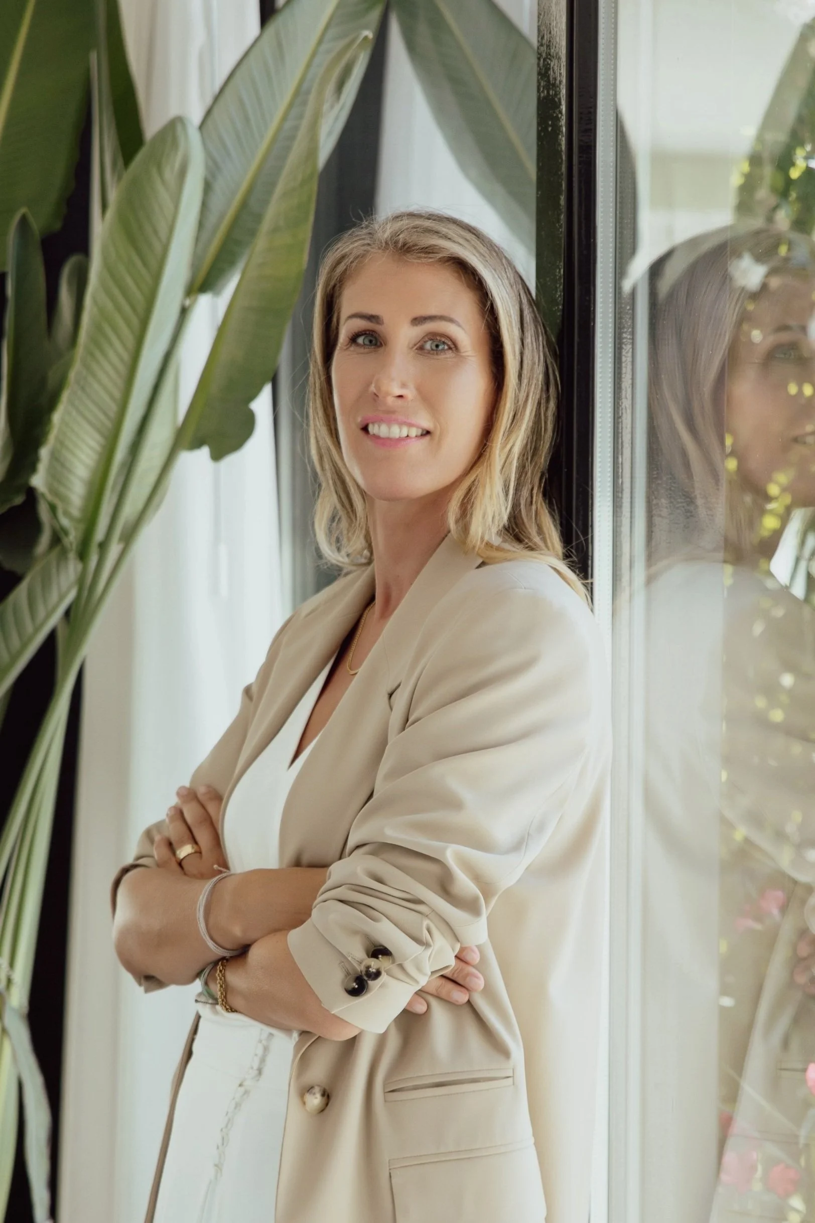 Confident female coach standing by sunlit window with lush greenery — representing grounded leadership, personal empowerment, and conscious presence.