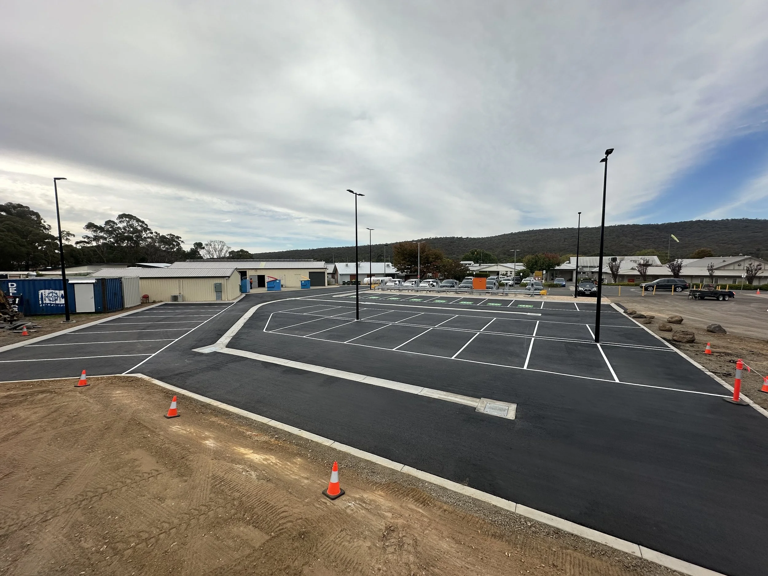 Newly paved parking lot with parking spaces and lamp posts, some orange safety cones, and a building with storage units in the background under cloudy sky.