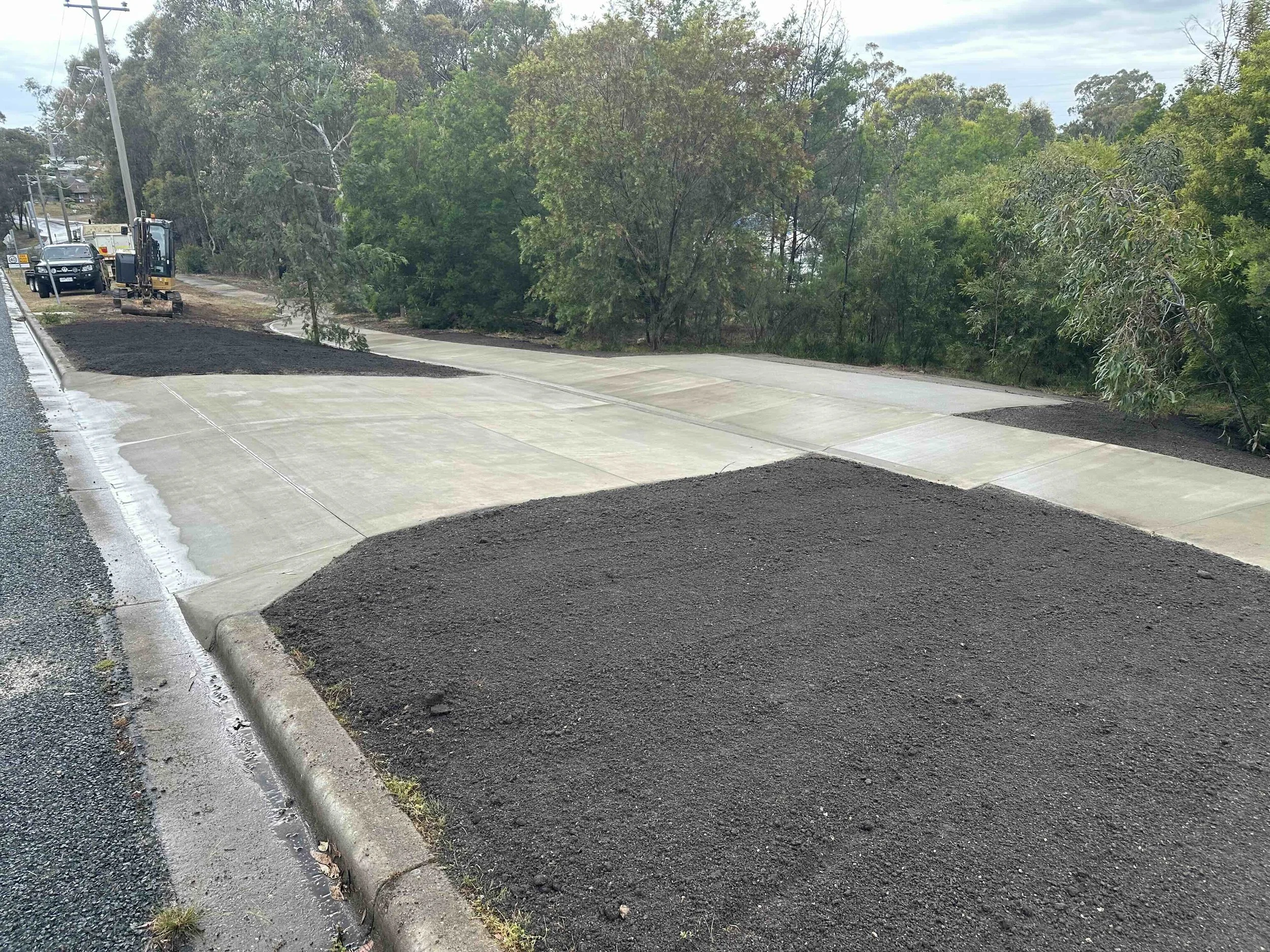Newly paved parking area with asphalt and concrete surfaces, surrounded by trees and bushes, with construction equipment and vehicles in the background.