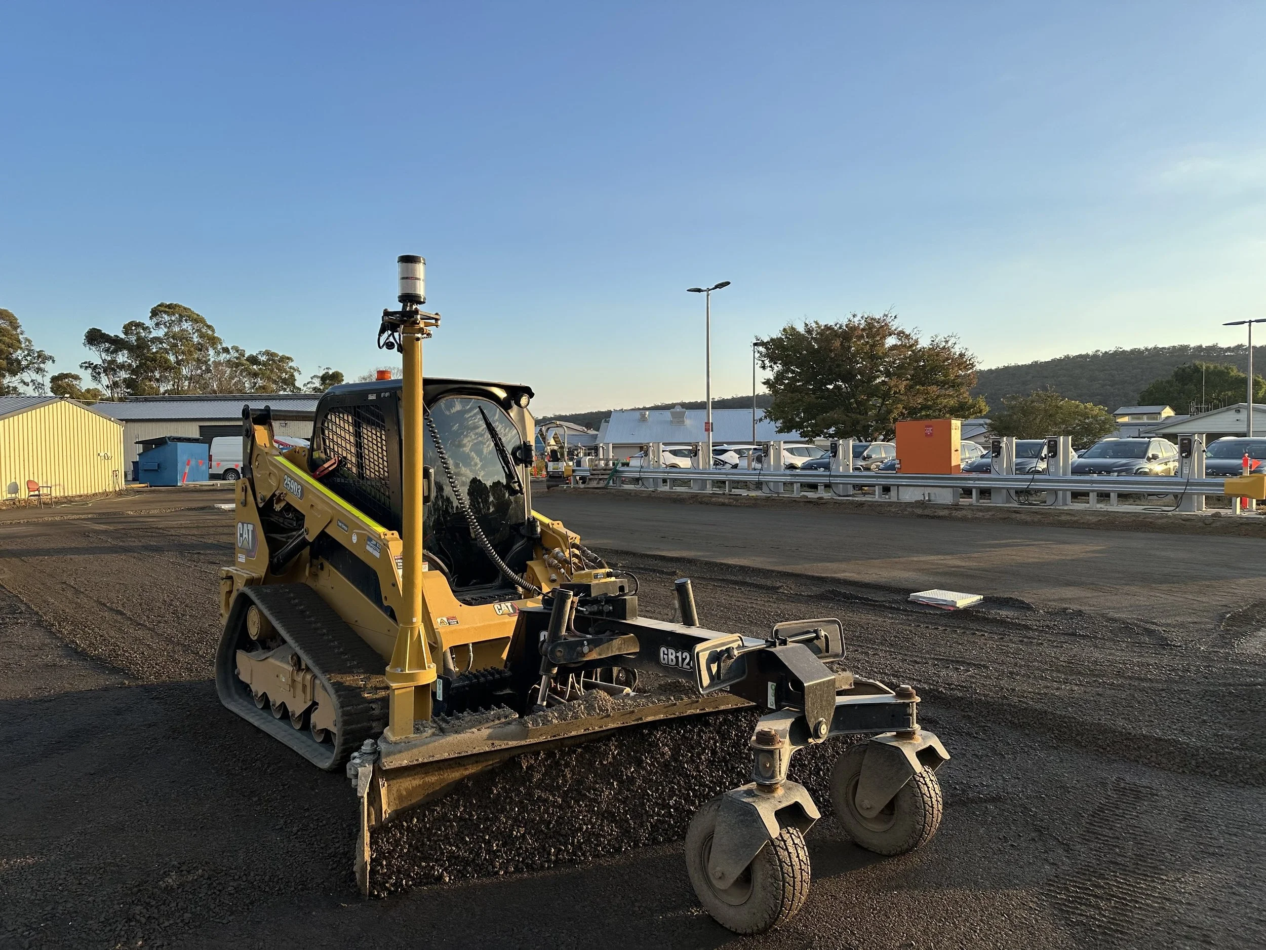 A construction vehicle on a dirt surface in a parking lot, with a background of trees, parked cars, and a building under a clear sky.