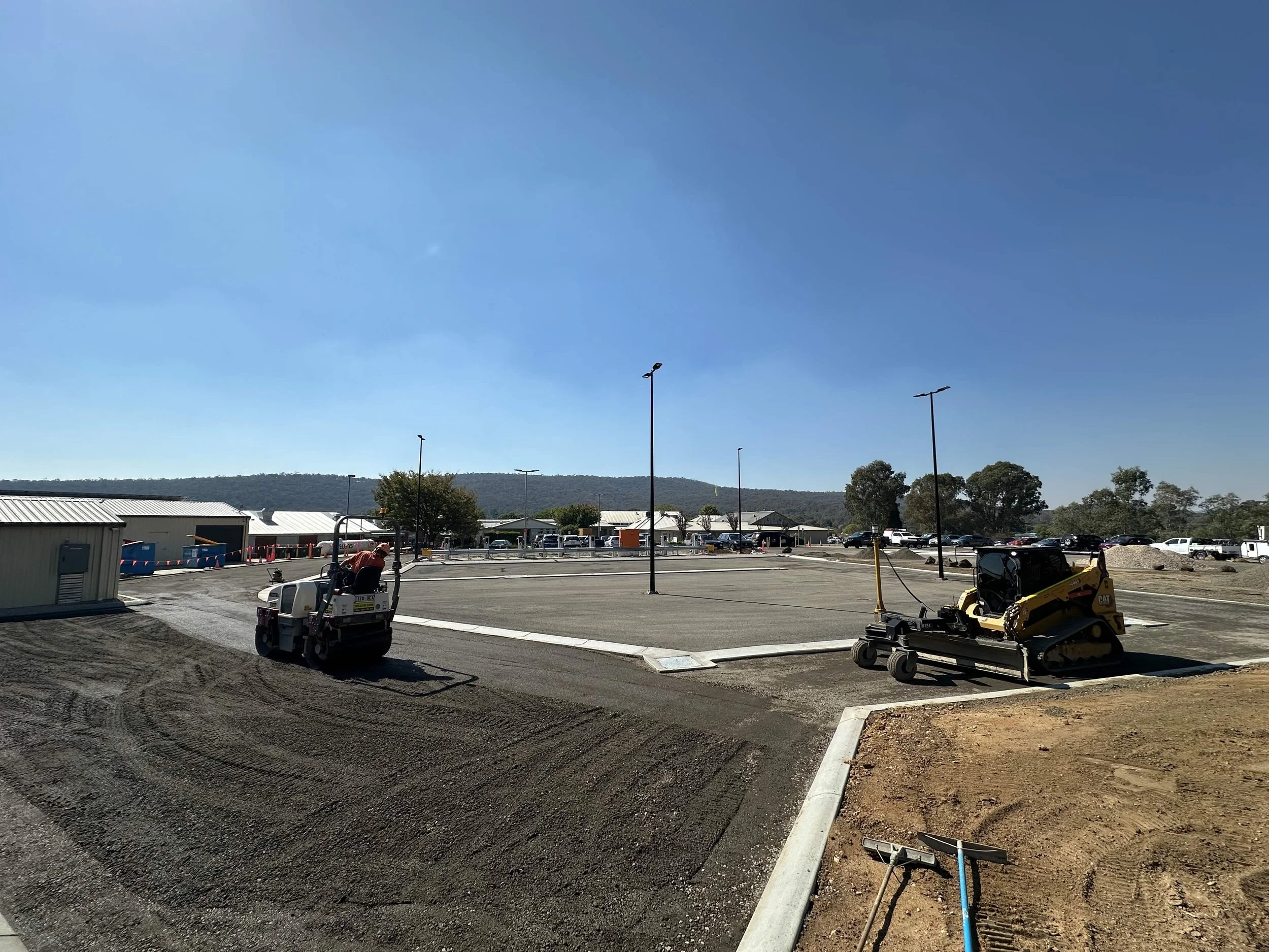 Construction site with paving equipment and an empty parking lot, with buildings and trees in the background under a clear blue sky.
