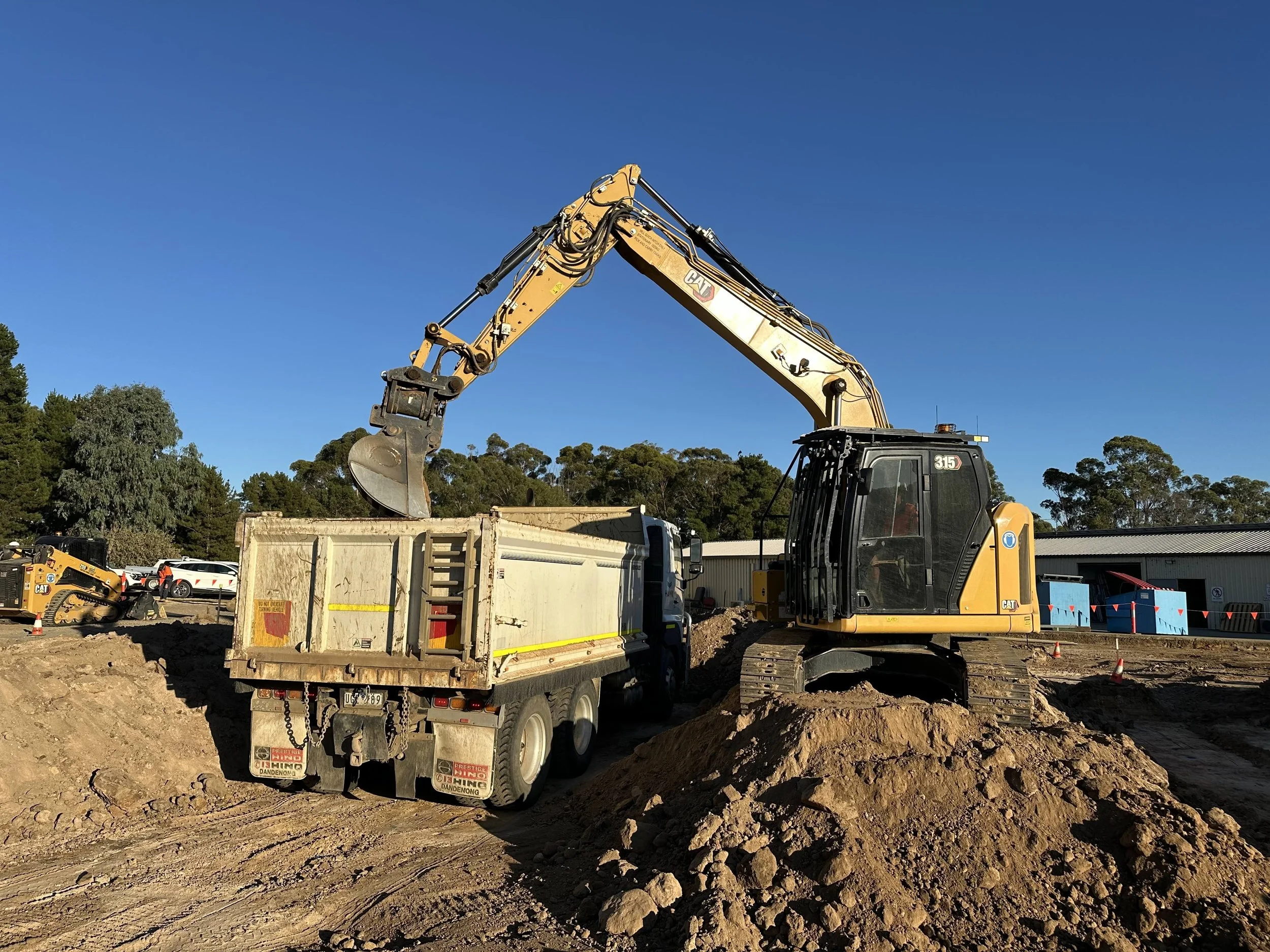 Construction site with a large yellow excavator loading dirt into a dump truck during daytime.
