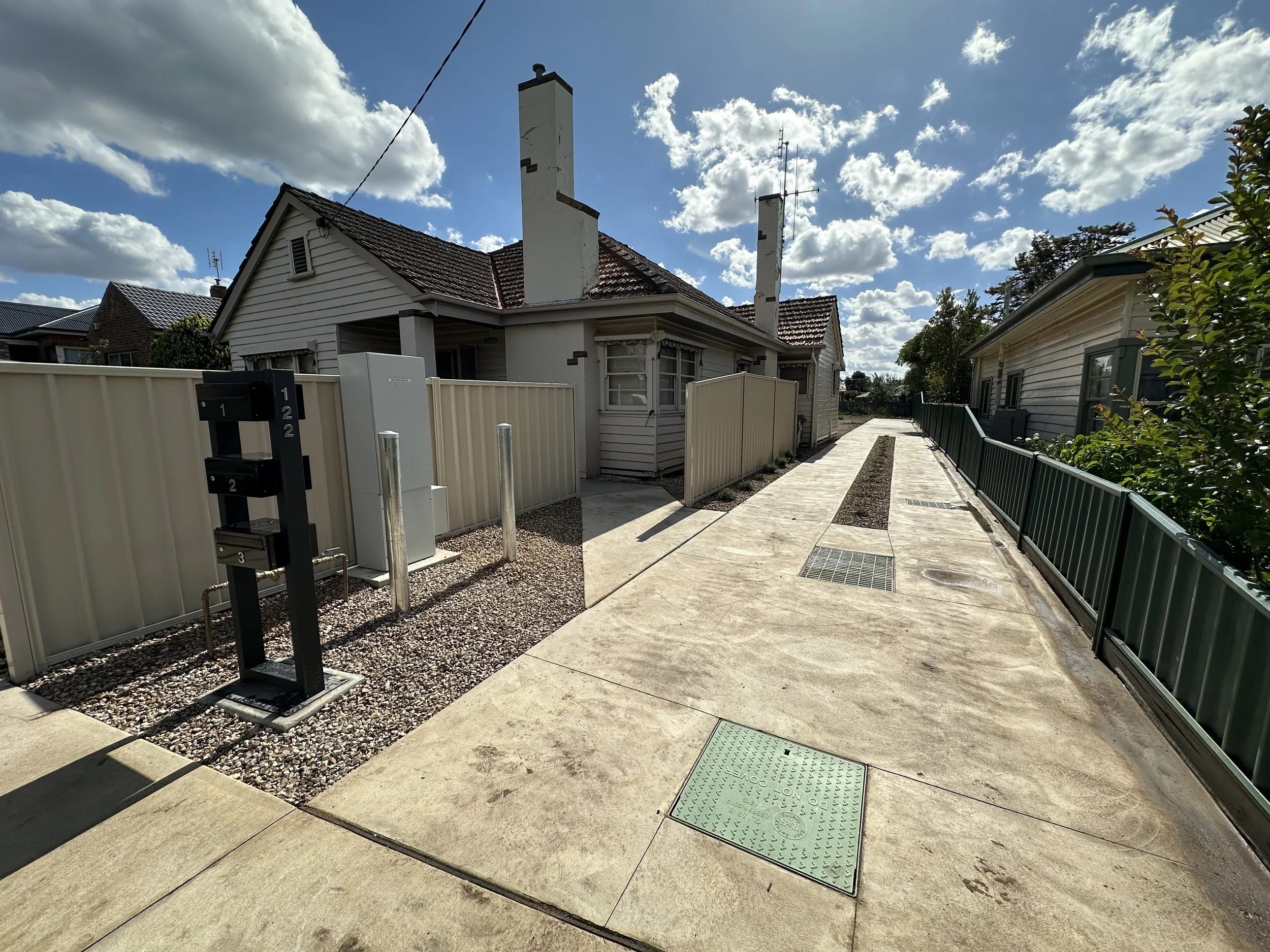 Residential alleyway with concrete sidewalk, green and beige fences, and utility boxes under bright daylight.