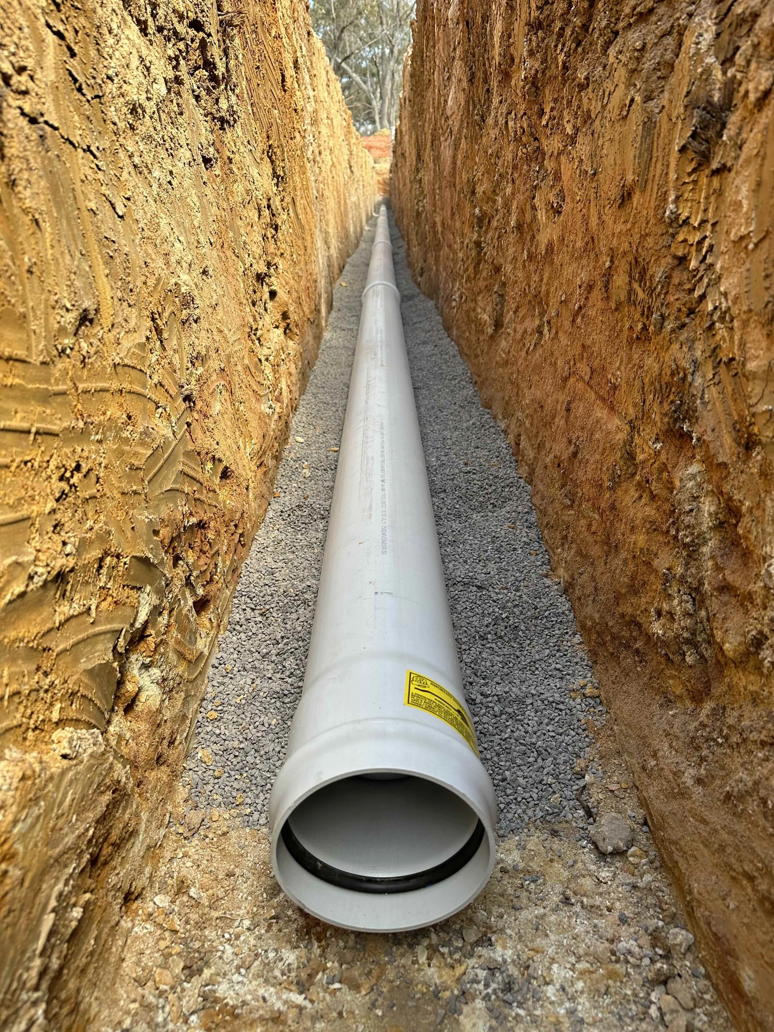 A large white pipe running through a narrow trench with exposed dirt walls on both sides.