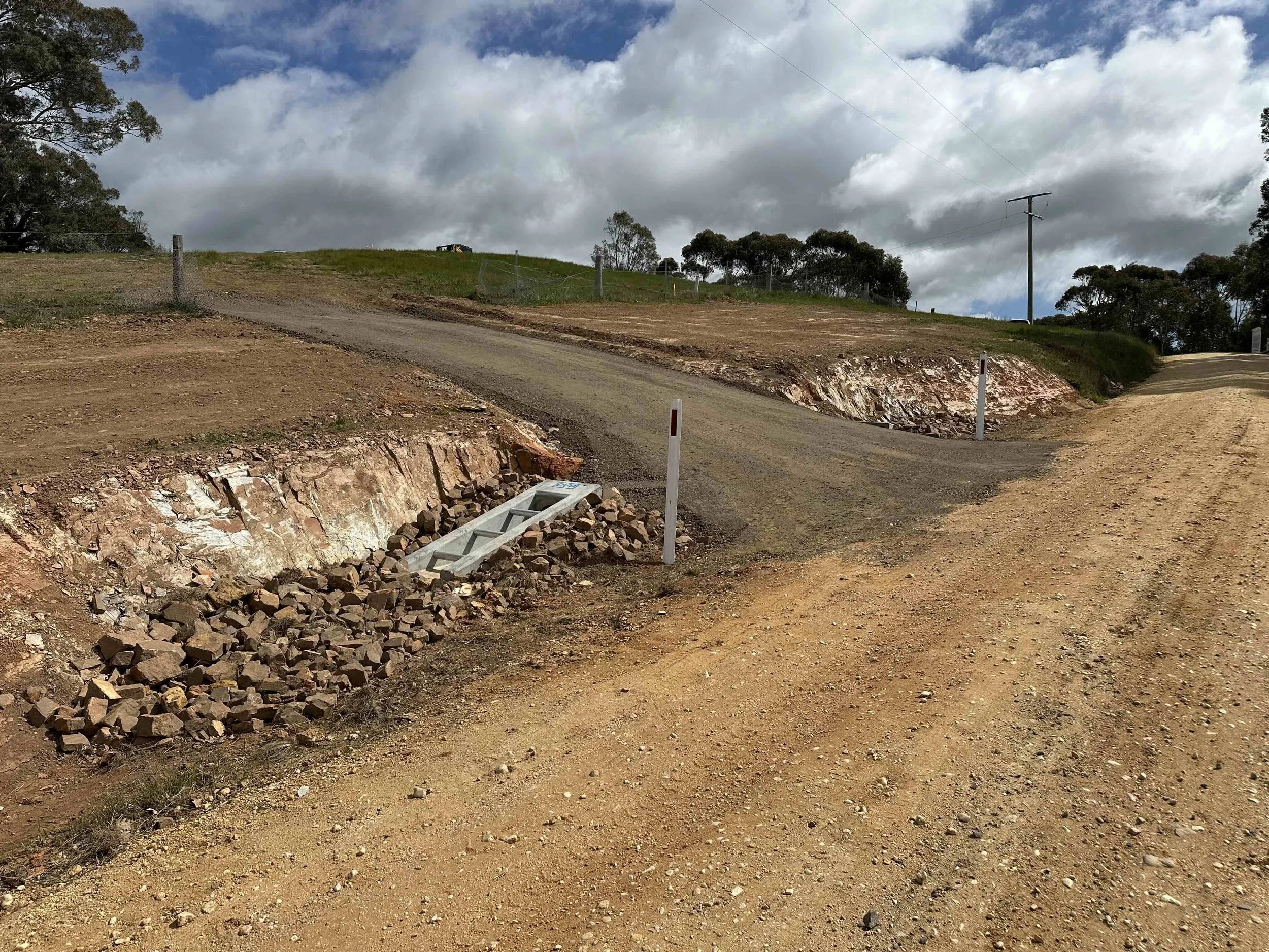 A dirt road with a section of broken pavement and a small hill in the background. There are some trees and power lines under a partly cloudy sky.