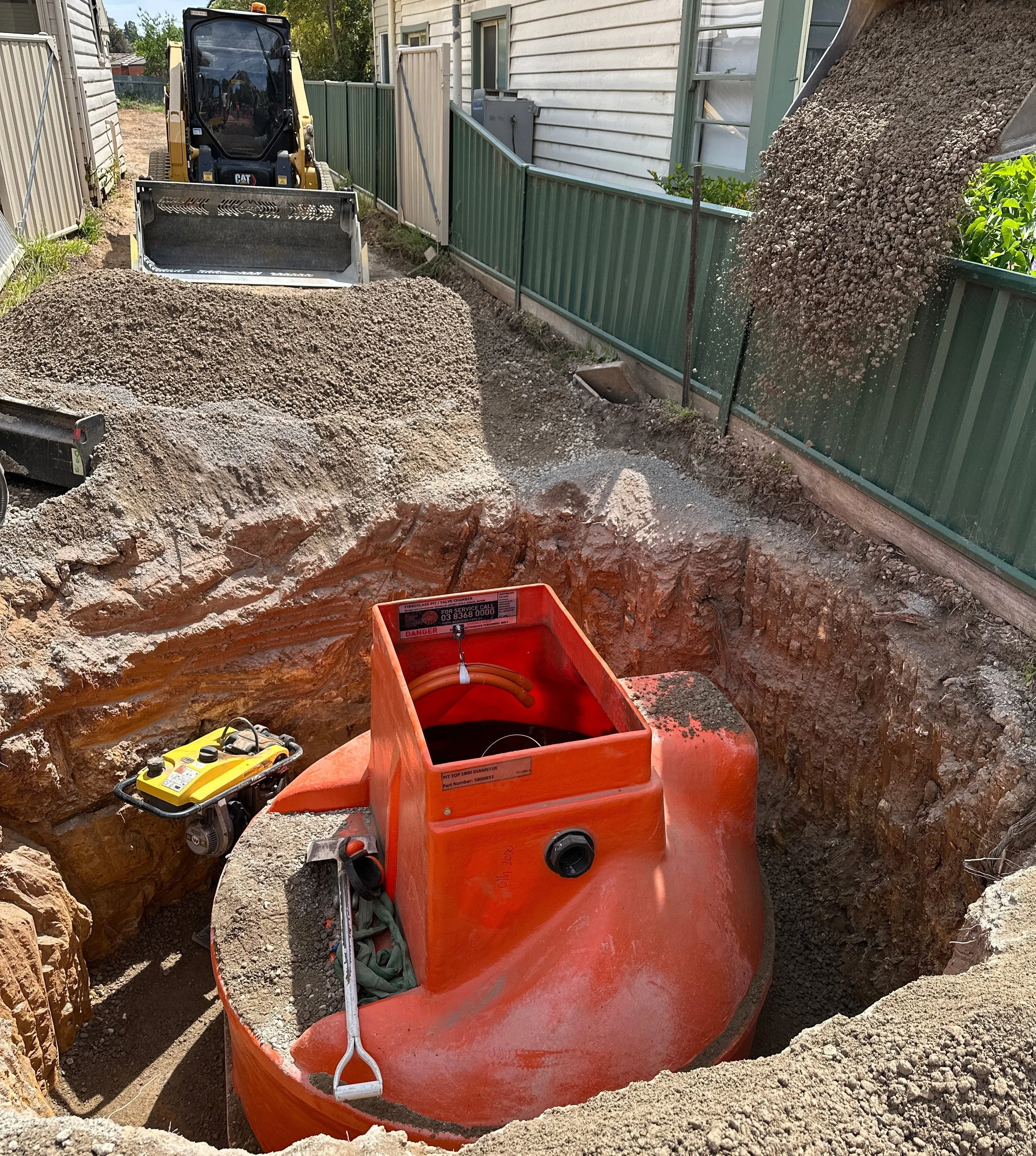 Construction site where a large excavation is taking place. A bright orange underground utility vault is being installed, with dirt and gravel around it. A small yellow portable generator is nearby, and a loader is depositing gravel into the excavation.