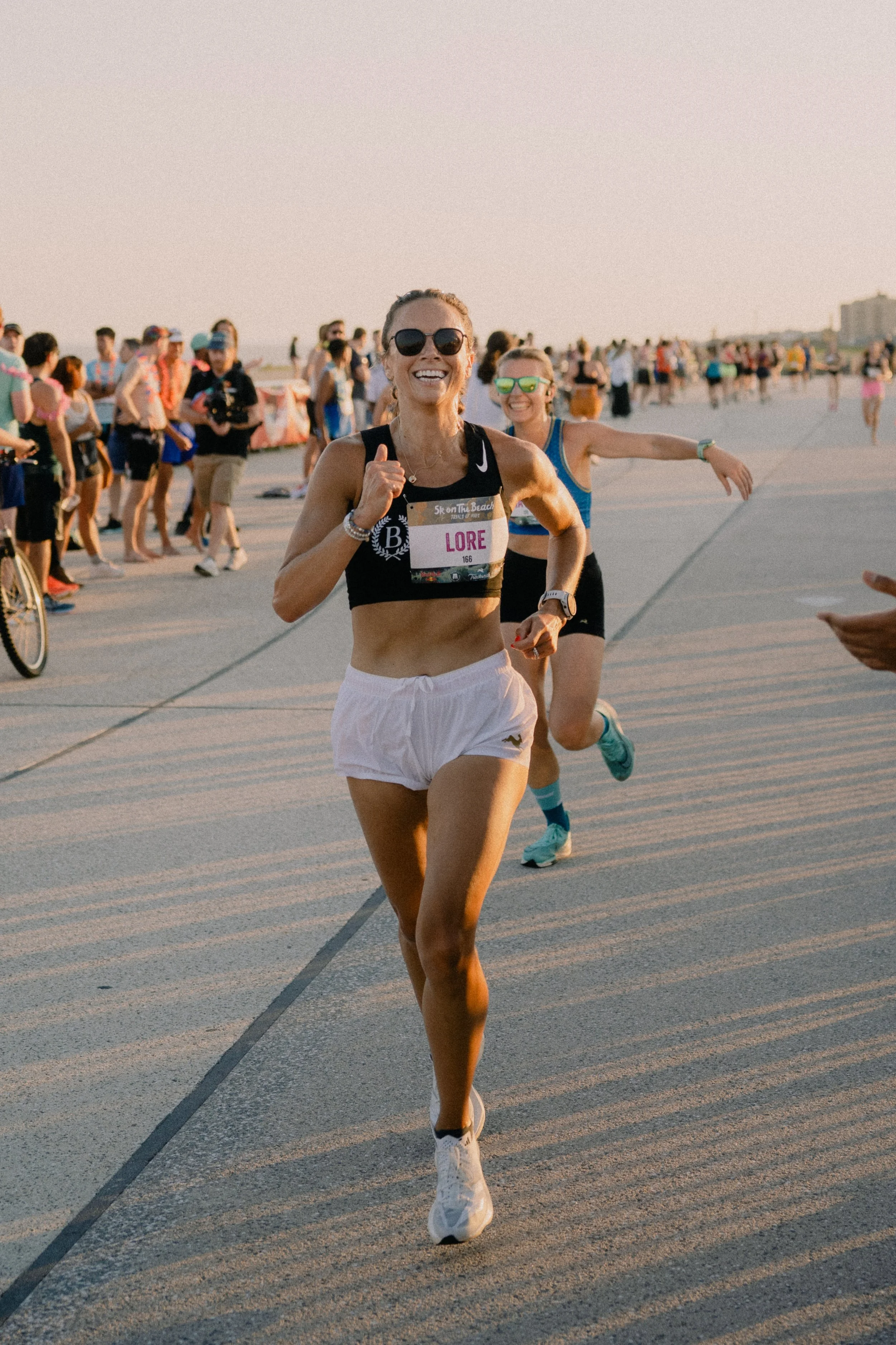 A woman running on a beach during a race or marathon, smiling and wearing sunglasses with a bib labeled "LORE". She is dressed in athletic clothing, and a crowd of people is visible in the background, also participating or spectating.