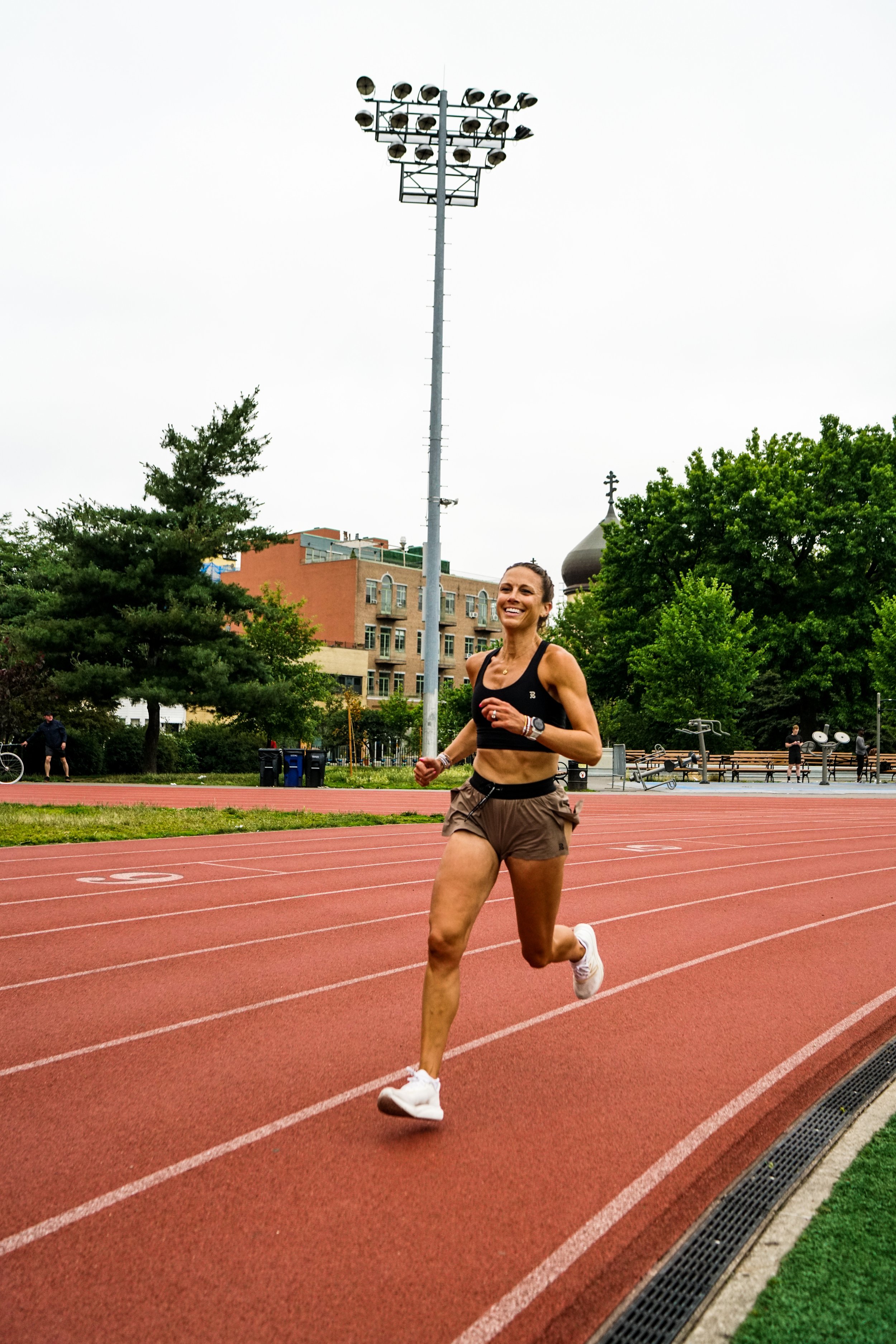 A woman running on a red track at an outdoor stadium, smiling. She is wearing a black sports bra, brown shorts, white running shoes, and a fitness watch. There are trees, buildings, and a tall stadium light pole in the background.