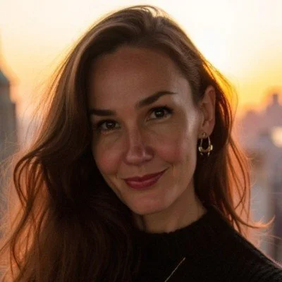 A woman with long brown hair smiling at sunset in an urban setting, wearing hoop earrings and a black top.