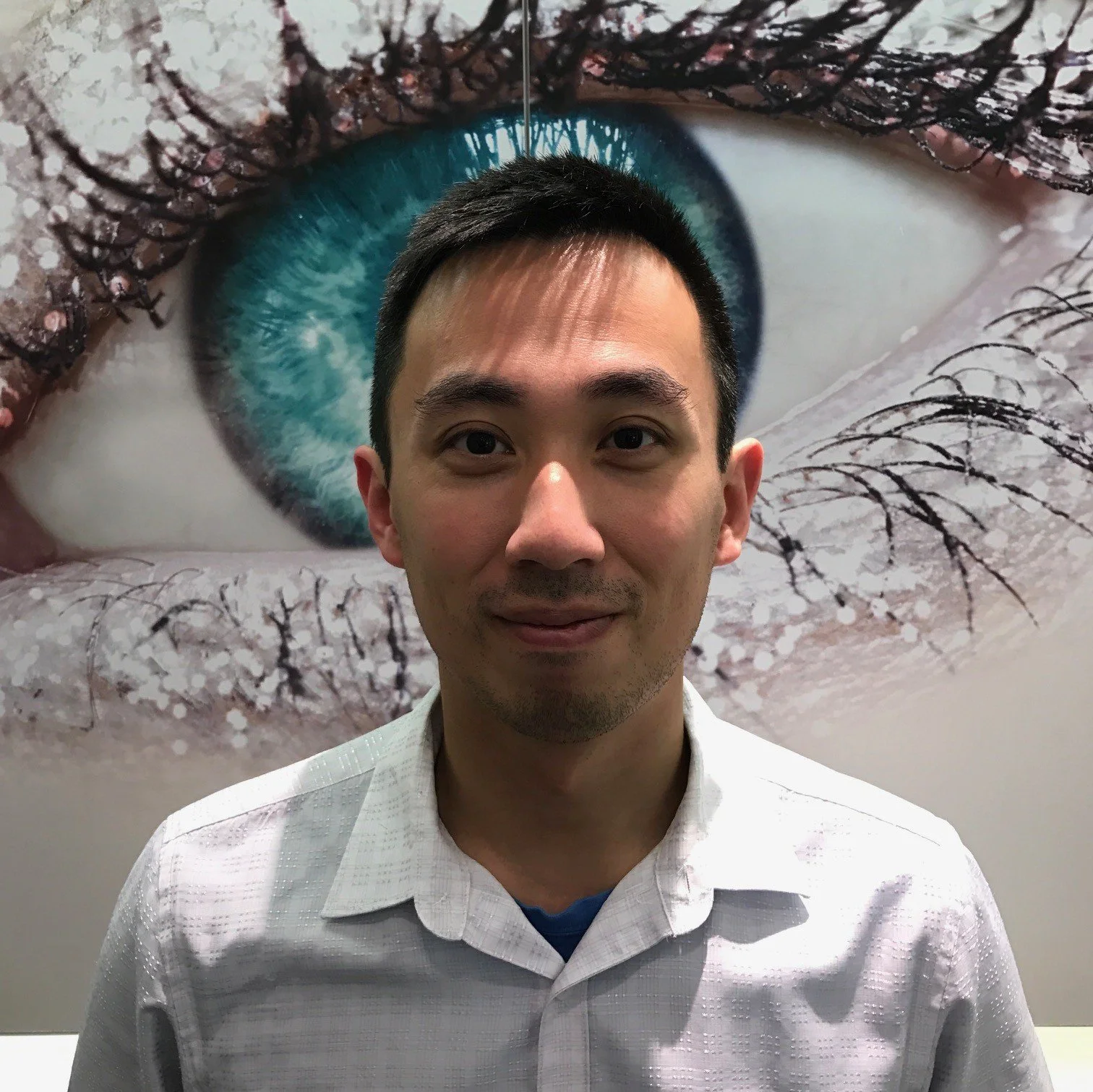 A man with short black hair wearing a white dress shirt standing in front of a large eye mural with a blue iris and detailed eyelashes.