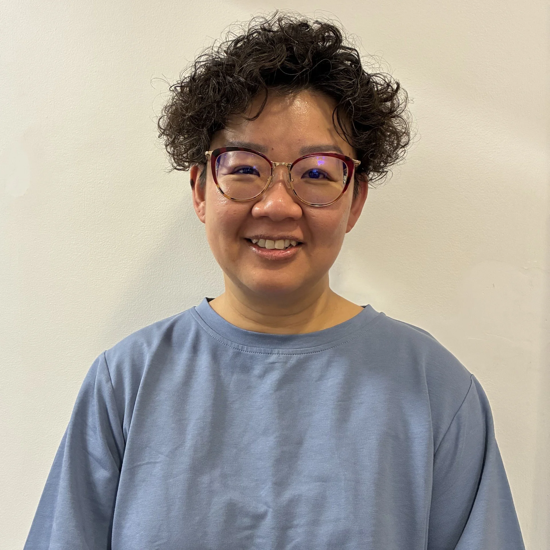 Close-up of a smiling person with curly dark hair, glasses, and wearing a blue shirt, standing against a plain light-colored wall.