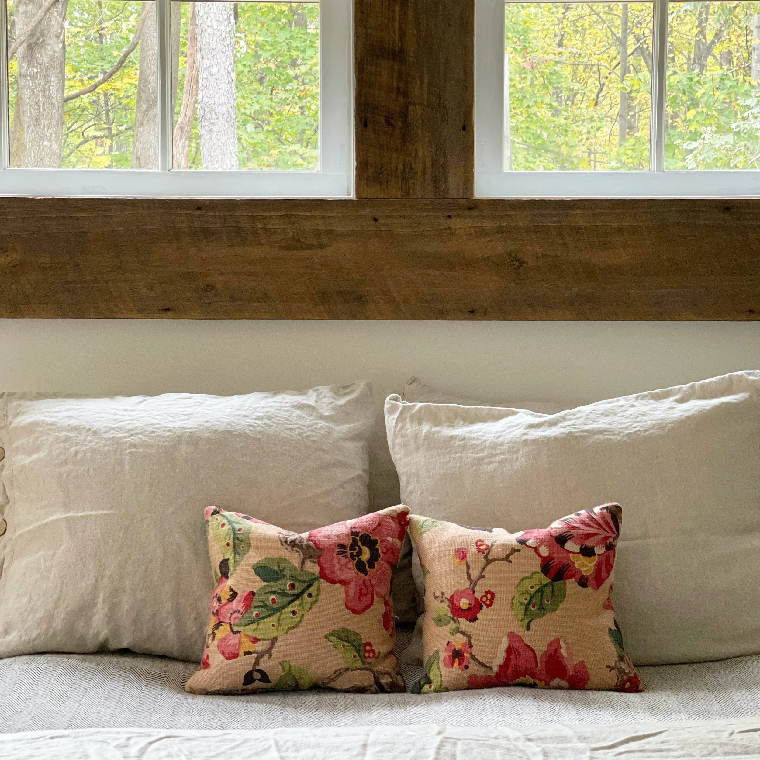 A cozy bed with two large white pillows and two smaller floral patterned cushions, set against a window with a view of trees and fall foliage.