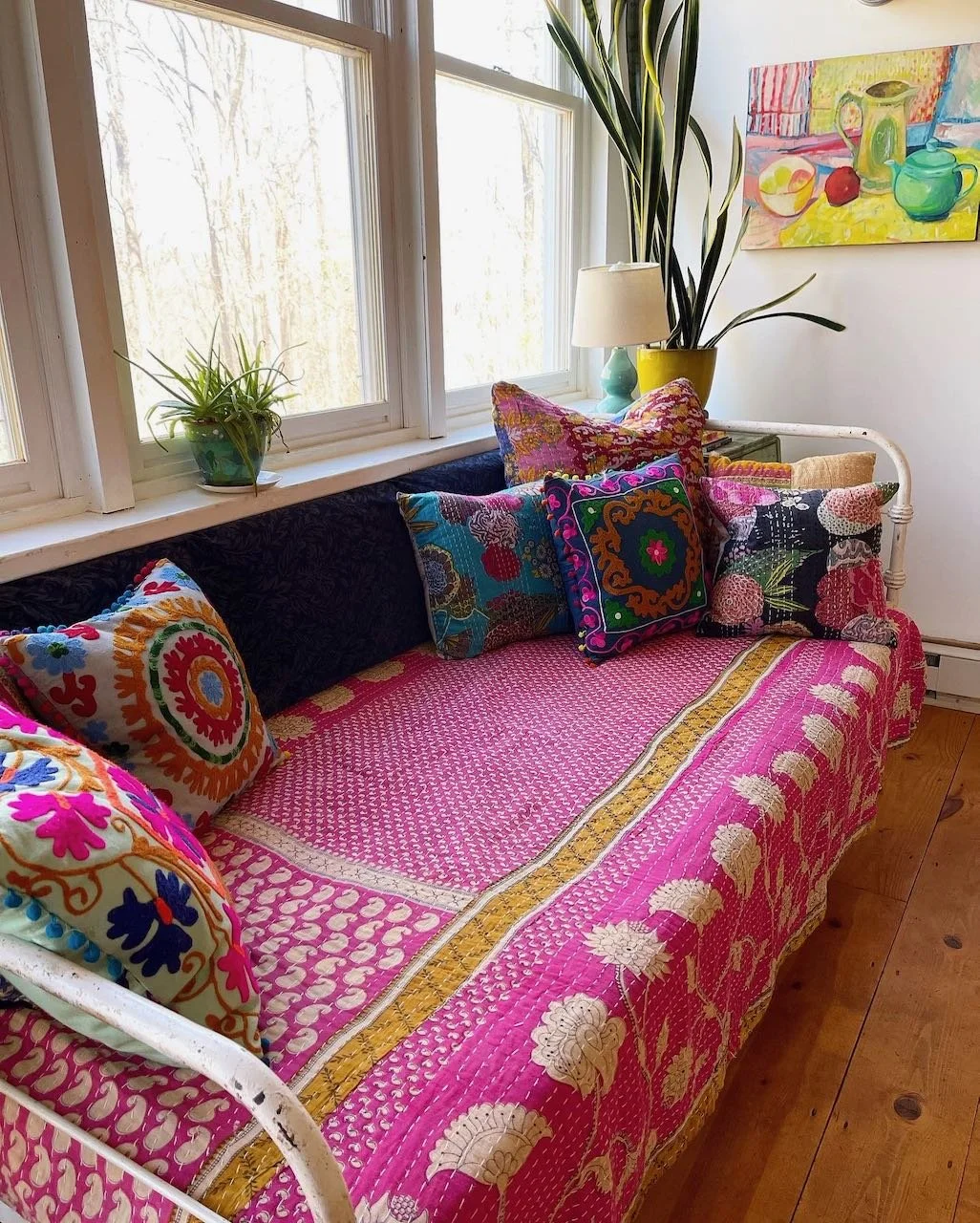 Colorful indoor space with a white vintage-style sofa covered in a bright pink quilt and multiple decorative pillows with vibrant embroidered and patterned designs. There is a large window behind the sofa with two potted plants on the windowsill, a yellow vase with a tall plant beside a table lamp, and a colorful still life painting of fruit and pitchers on the wall.