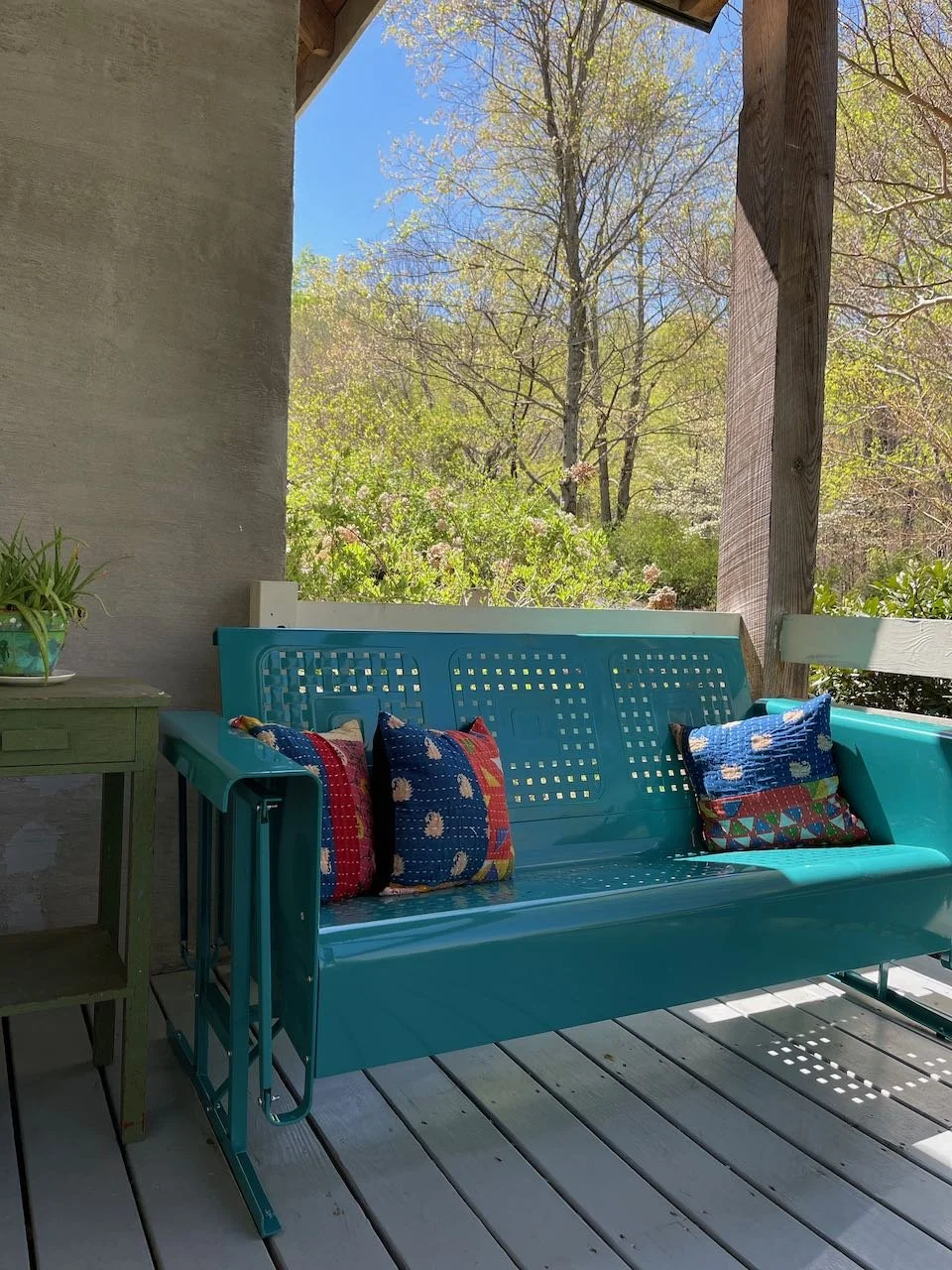 A porch with a blue metal bench with colorful patterned pillows, a small green table with a potted plant, surrounded by trees with green leaves and a clear blue sky.