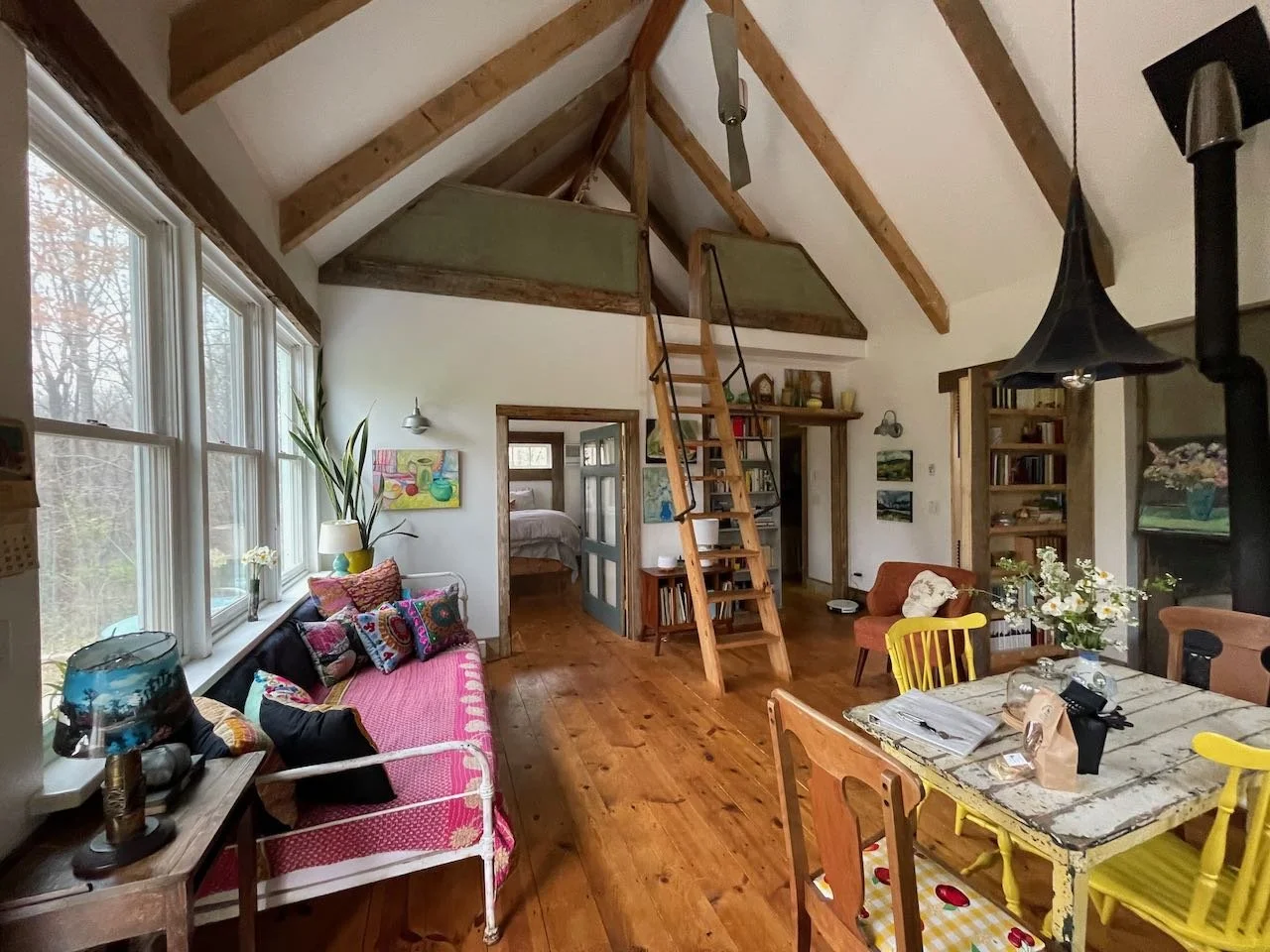 A cozy living room with a vaulted ceiling featuring wooden beams, large windows letting in natural light, a pink and patterned sofa, a rustic dining table with mismatched chairs, and a loft area accessible by a wooden ladder.