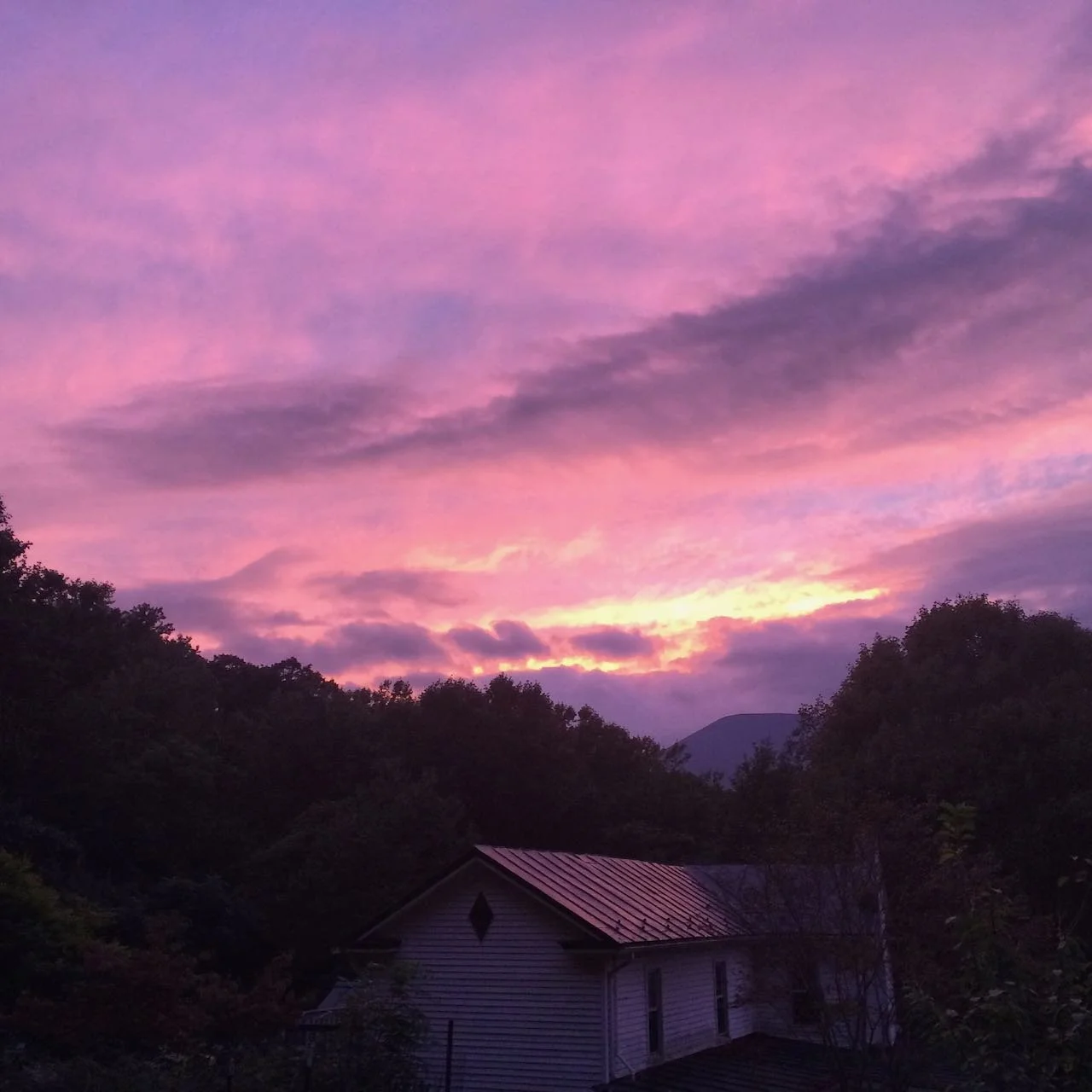 Sunset sky with pink and purple clouds over a rural landscape with trees and a house.