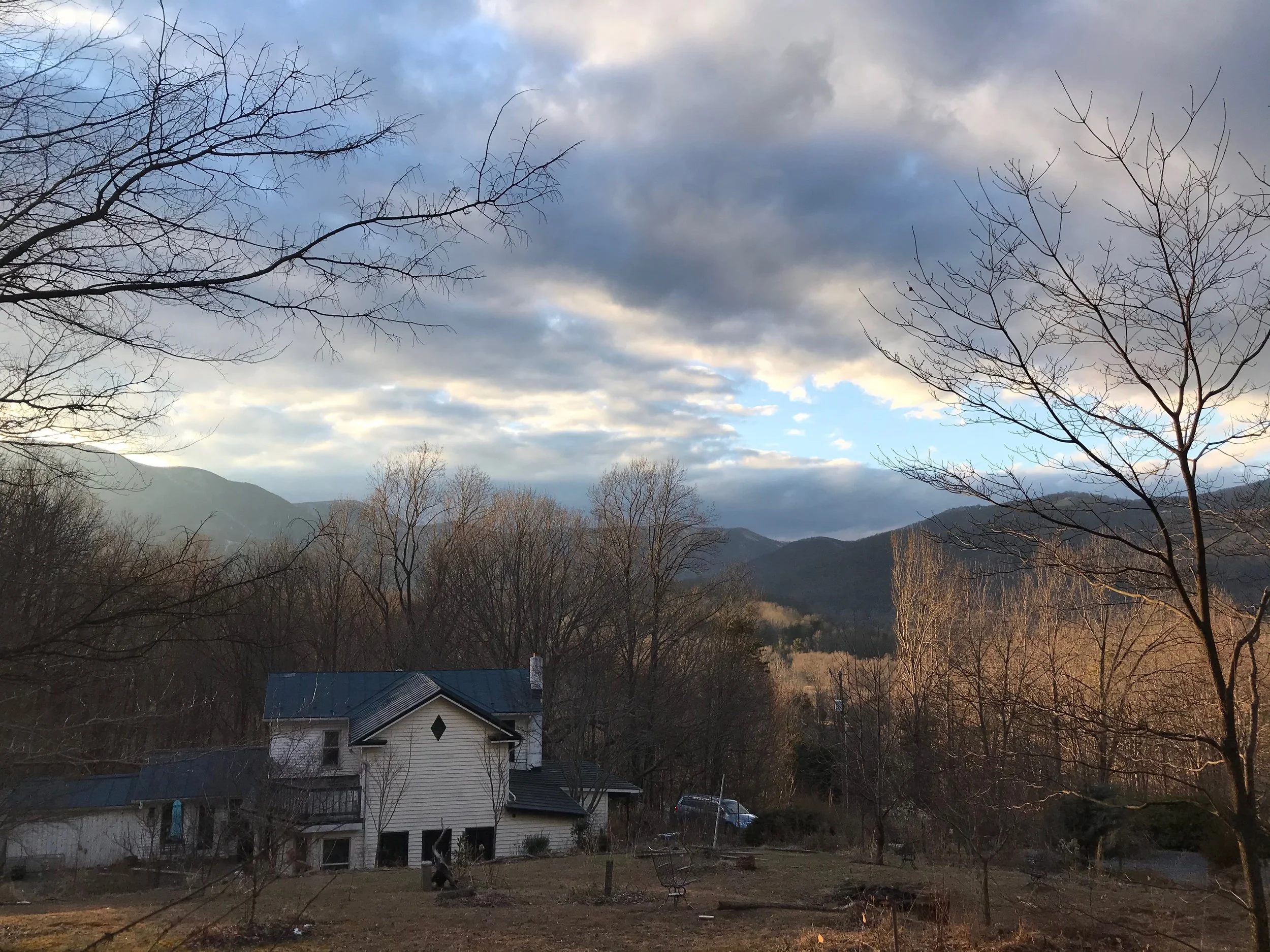 View of a house and yard with mountains and cloudy sky in the background, leafless trees in late fall or winter.