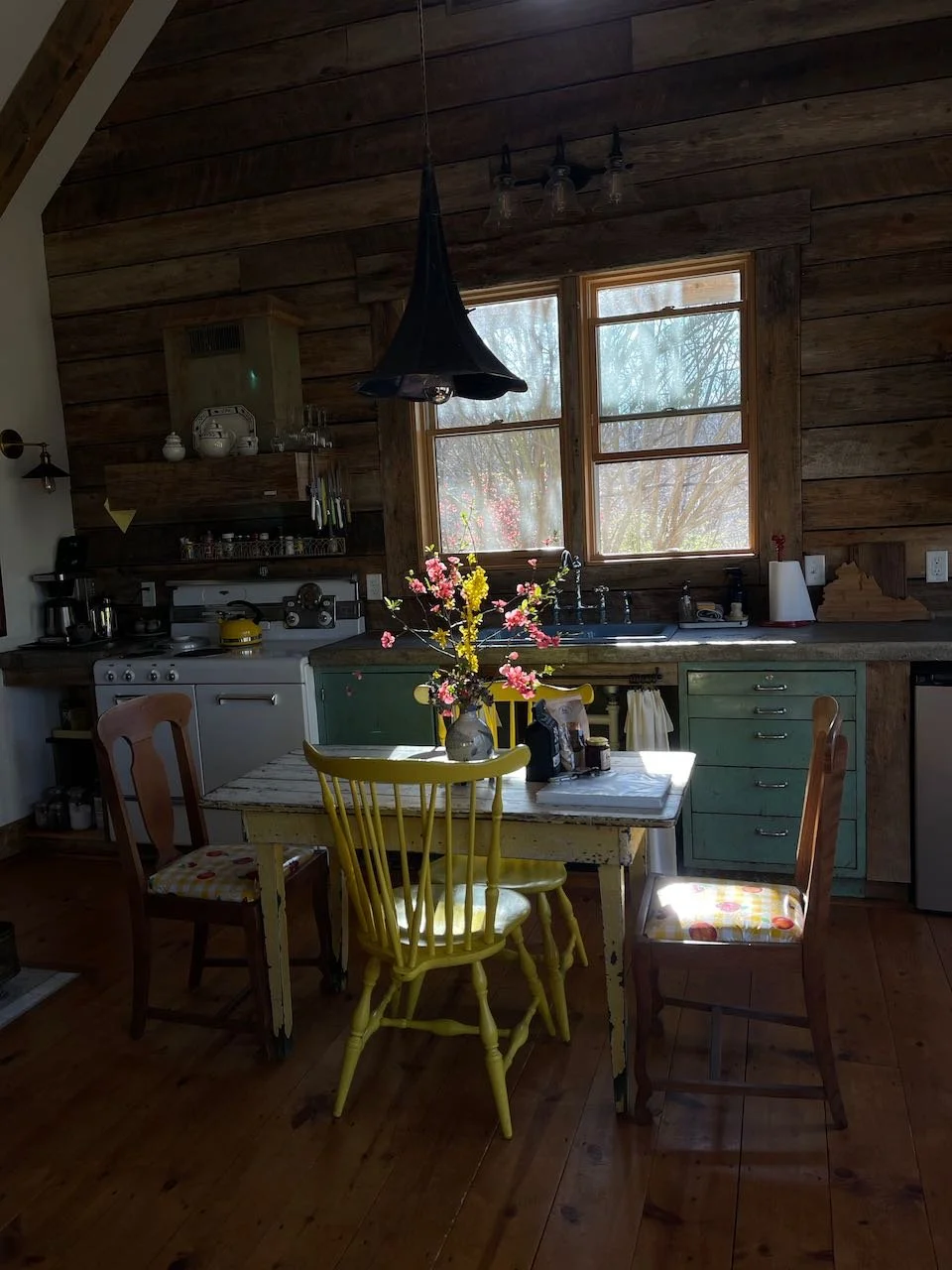 A rustic kitchen with natural wood walls and ceiling, featuring a small dining table with a vase of pink and yellow flowers, surrounded by mismatched chairs. Two windows above a green countertop let in natural light. Kitchen appliances and décor are visible, including a stove, shelves, and a hanging black pendant light.