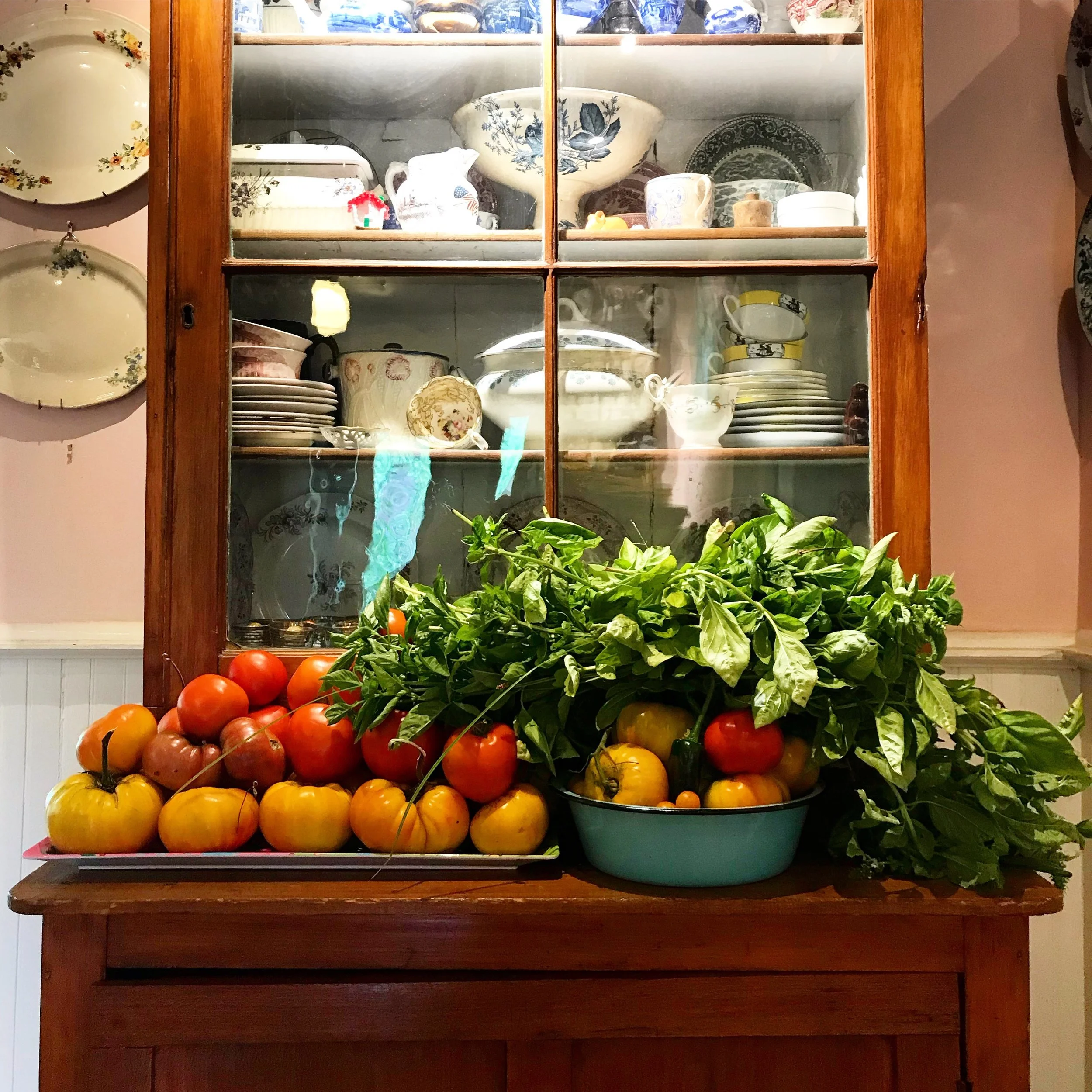 A wooden table with heirloom tomatoes, one with basil, placed in a tray and a bowl. Behind the table is a wooden cabinet with glass doors displaying dishes, bowls, and cups.