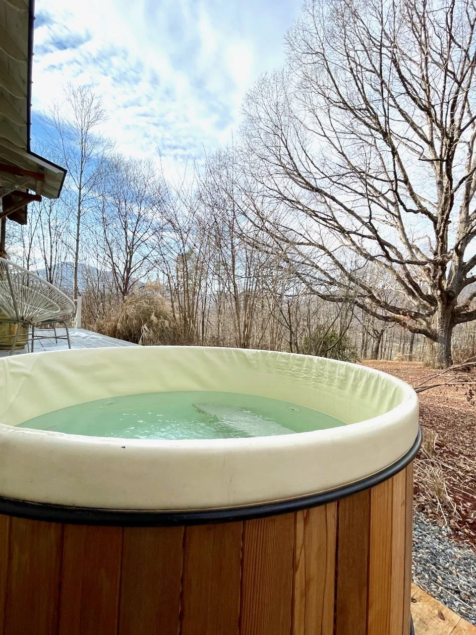 A hot tub with water on a wooden deck outside near leafless trees under a partly cloudy sky.