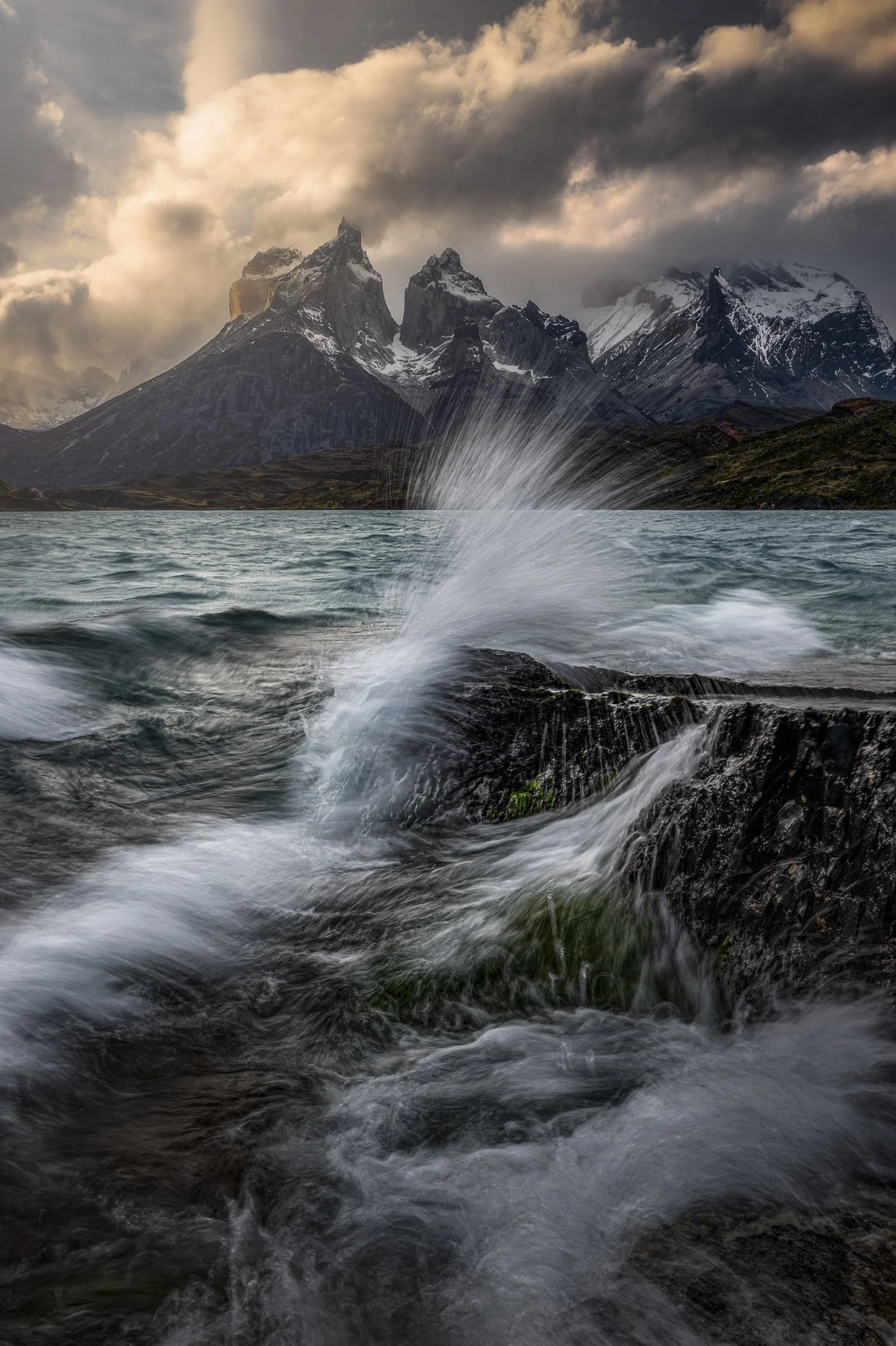 Mountain range with snow peaks behind a body of water, with ripples and splashes from water contact with rocks in the foreground, dark and cloudy sky overhead.