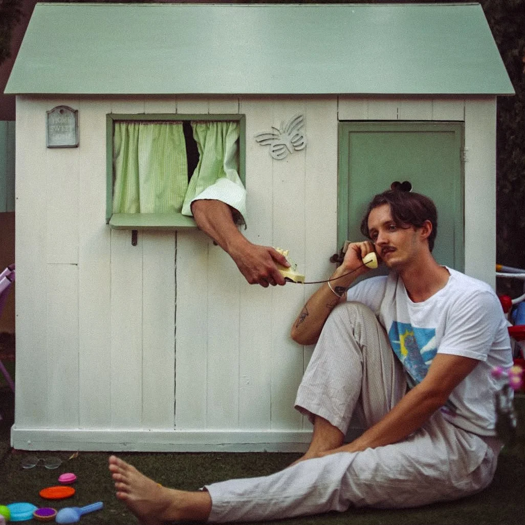 A person sitting outside a small wooden playhouse receiving a phone call while a hand extends out of the playhouse holding a remote control toward the person. The person has short brown hair, a mustache, and is wearing a white t-shirt and light-colored pants. The playhouse has a green roof, a small window with green curtains, a butterfly decoration, and a green door. There are scattered colorful bottle caps on the ground.