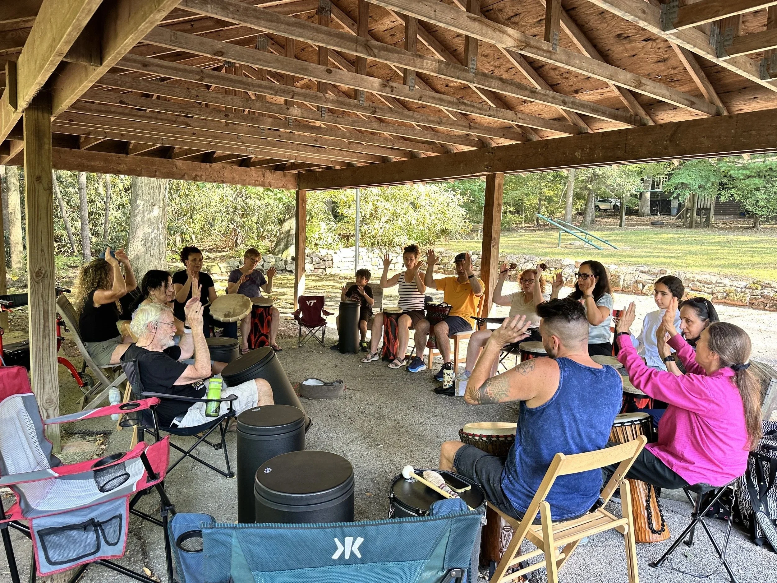 Dani Fiore is facilitating a group of drummers at an outdoor pavilion in the summer.