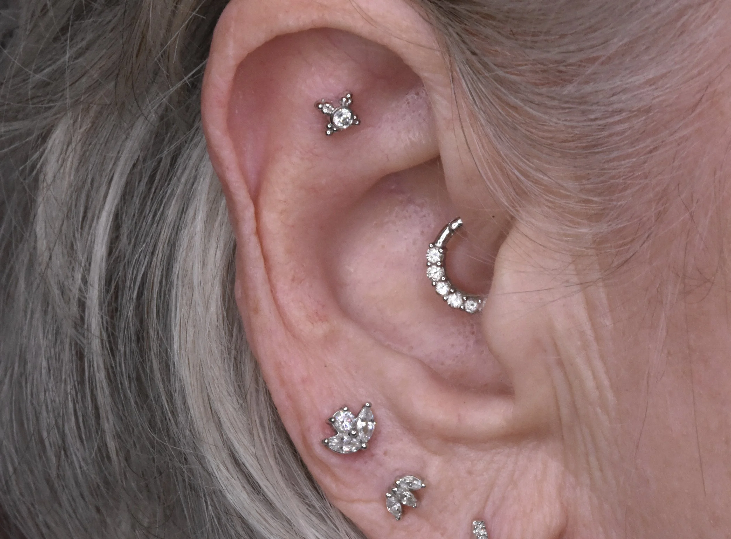 Close-up of a person's ear with multiple piercings, adorned with various silver earrings featuring clear gemstones, including a helix piercing, a conch piercing, a tragus piercing, and a lobe piercing with different decorative designs.