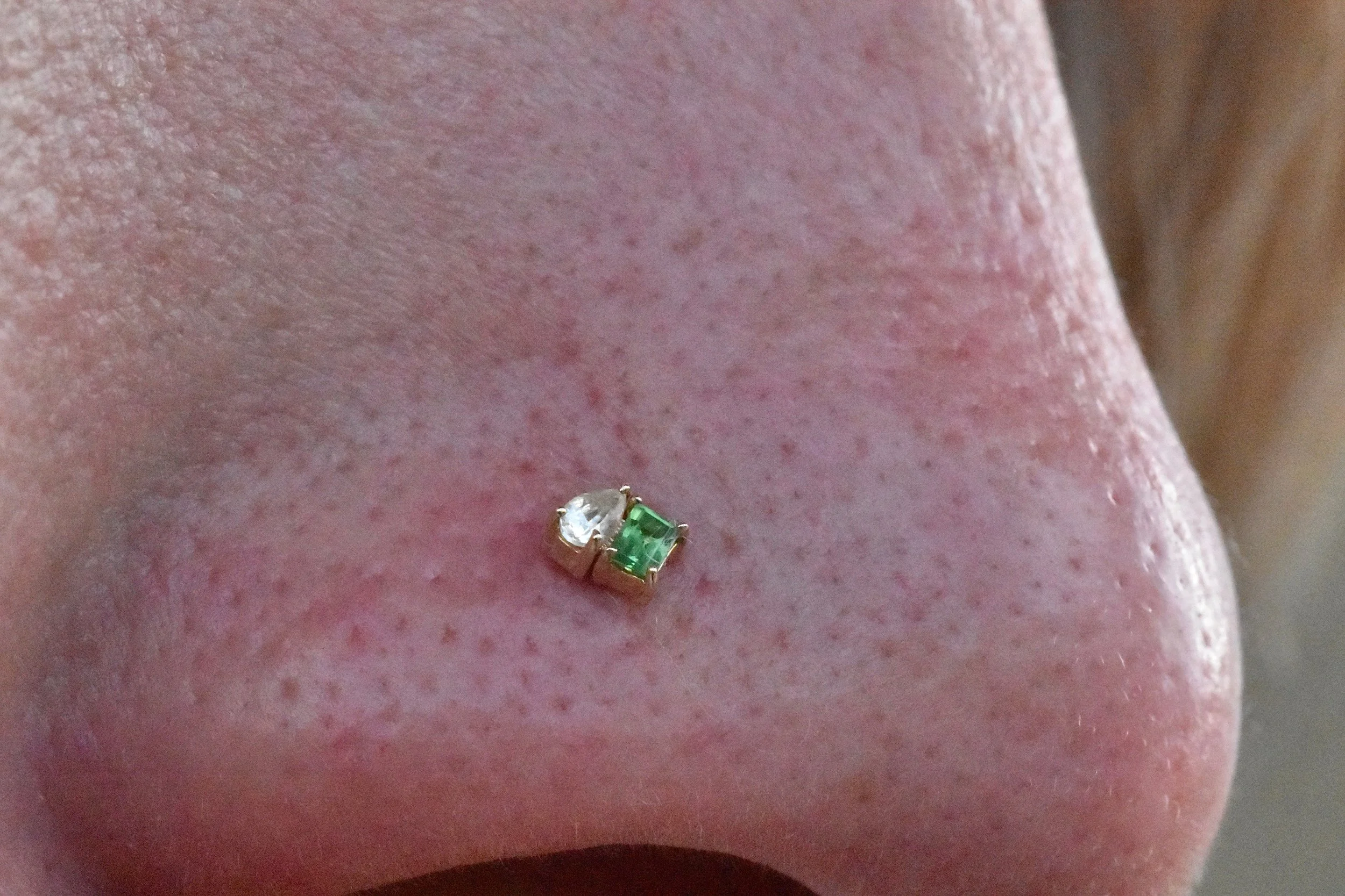 Close-up of a nose with a decorative nose ring featuring a large white gemstone and a smaller green gemstone.