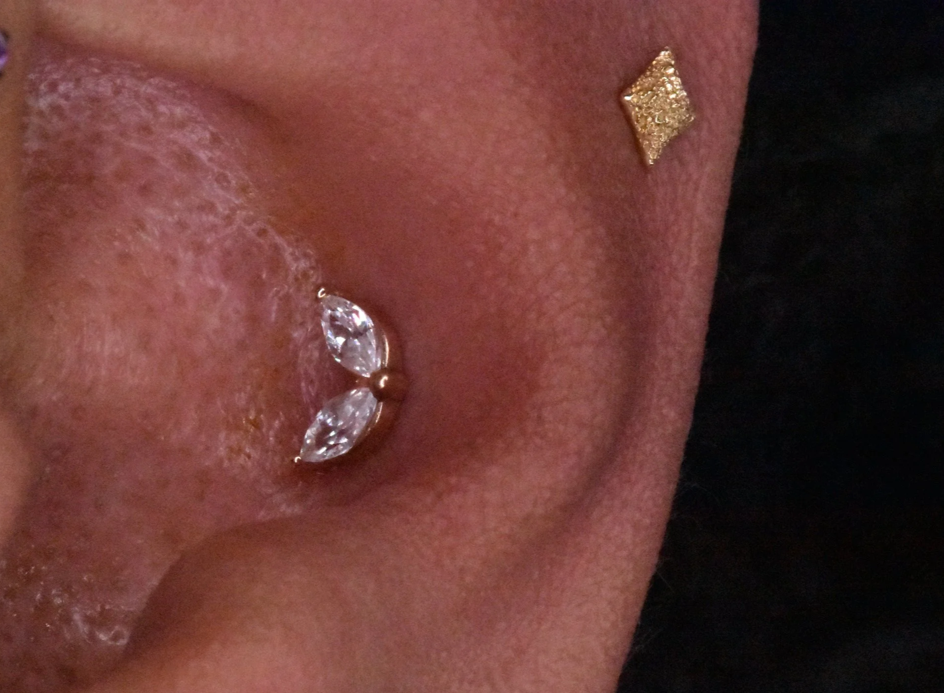 Close-up of a person's face, showing part of their nose and mouth, with jewelry including a gold square-shaped stud earring and a nose piercing with a large clear gemstone.