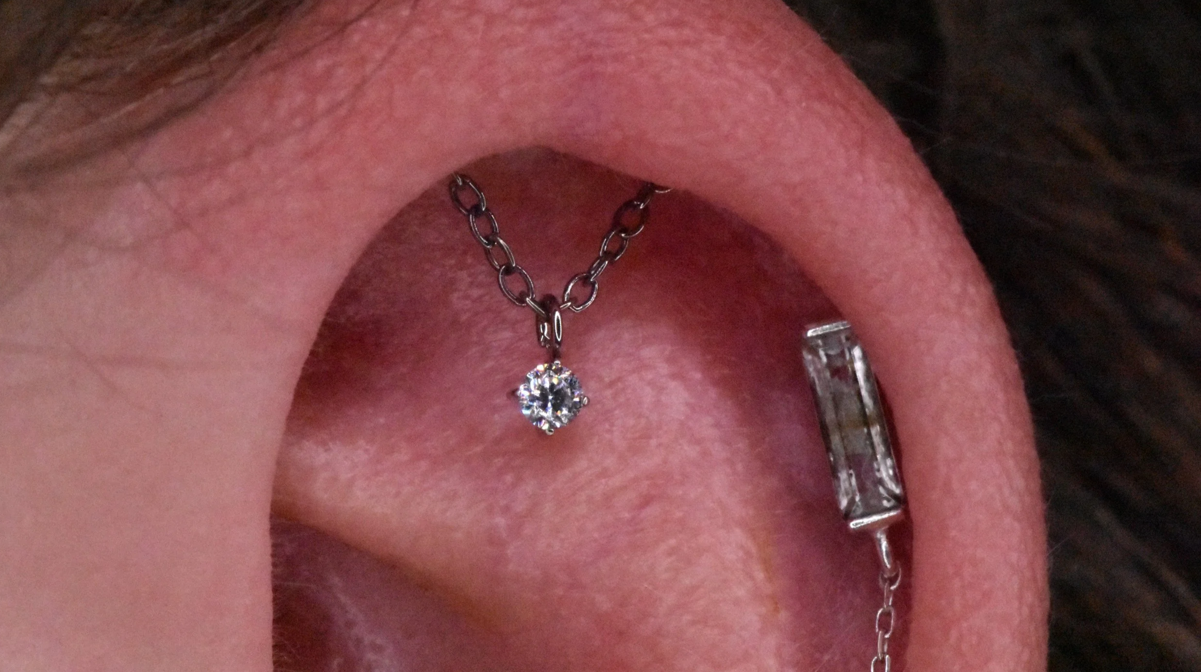 Close-up of a person's open mouth showing a tongue piercing with a chain and a diamond pendant, with visible pink tongue and surrounding skin.