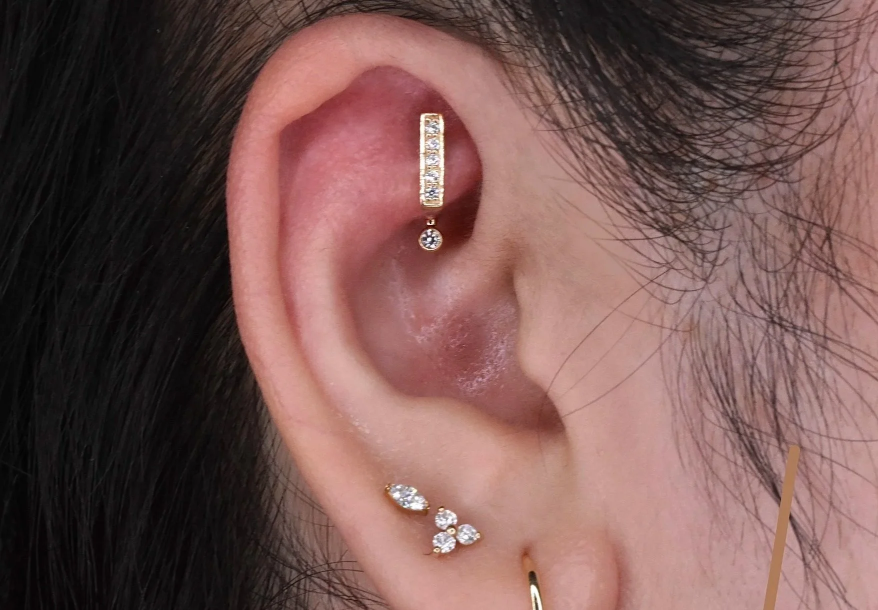 Close-up of a person's ear decorated with multiple gold earrings, including a vertical row of small crystals, a dangling crystal, and additional smaller earrings with crystals near the earlobe.
