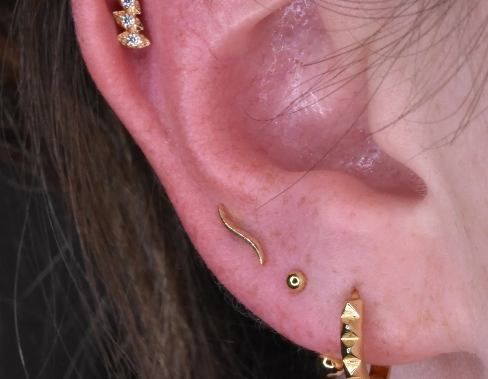 Close-up of a person's ear pierced with multiple gold earrings, including a curved barbell, a small stud, and a triangle-shaped earring, with visible skin and hair.