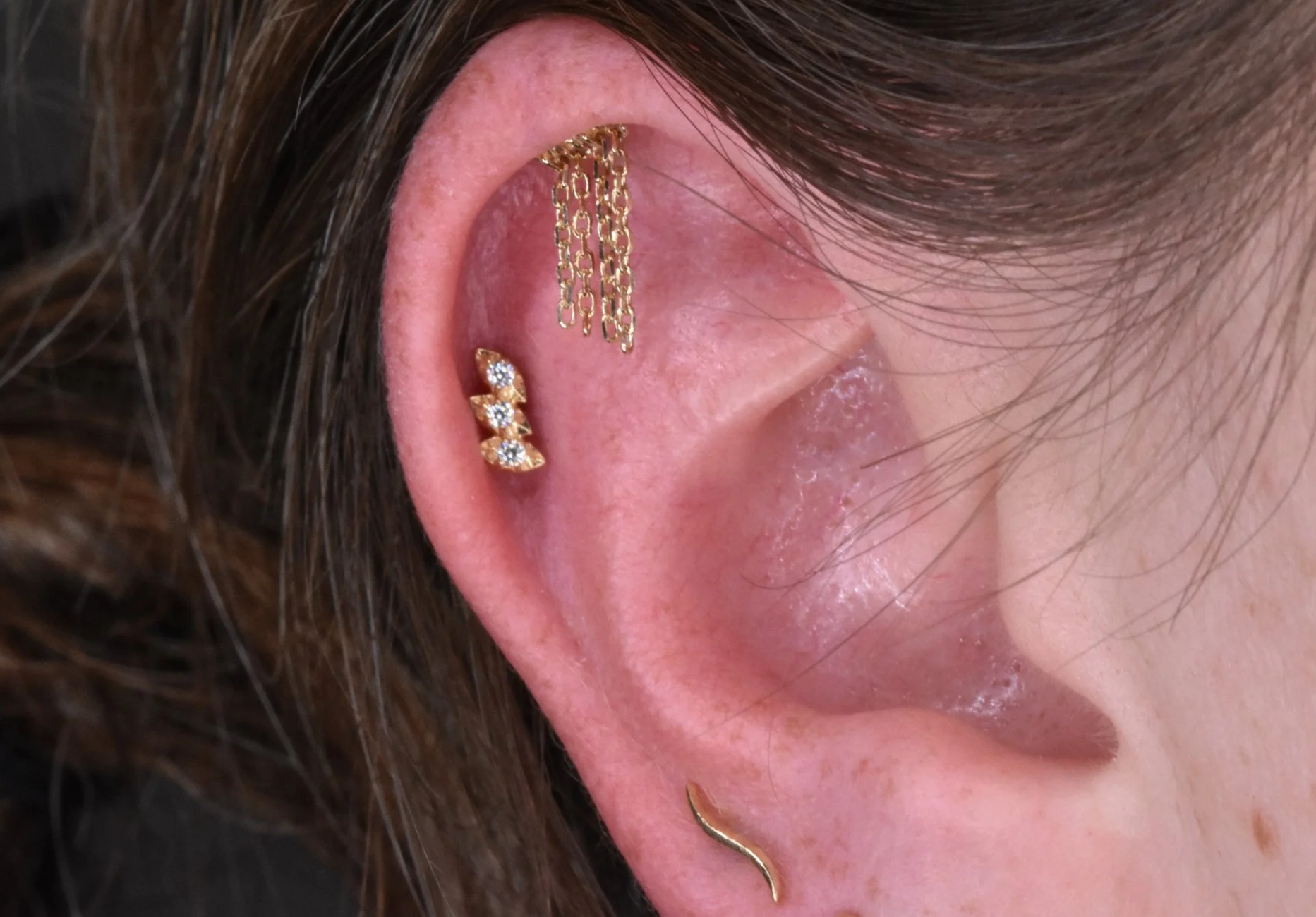 Close-up of a person's ear with multiple piercings, including a gold chain ear cuff, a gold earring with three diamonds, and a gold helix earring, with brown hair surrounding the ear.